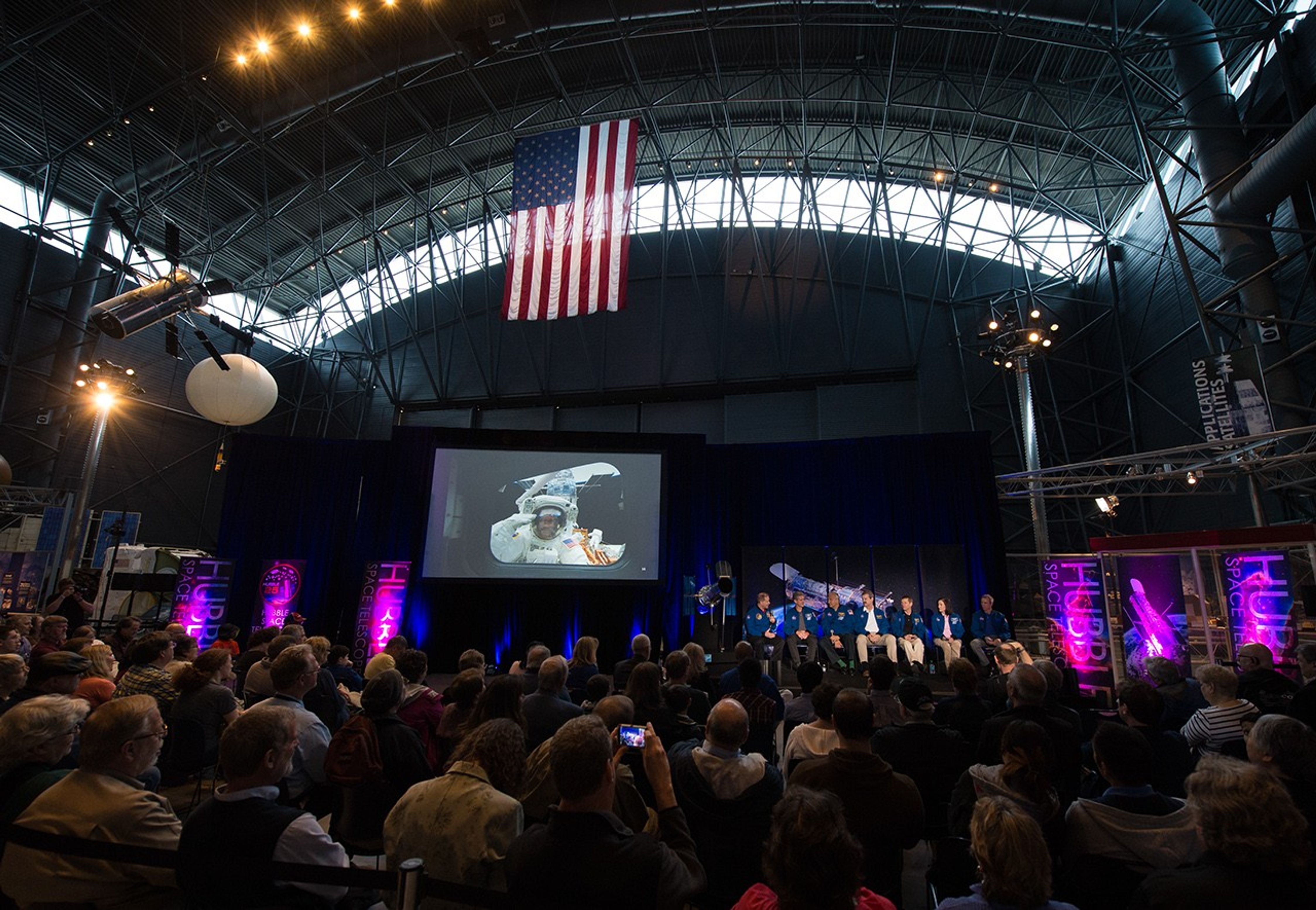 An image of the astronaut panel onstage taken from farther back in the audience. The audience members listen attentively, and some take pictures with their phone. To the left of the astronauts is the Hubble model, and to the left of that, a large screen displaying images from the final Hubble servicing mission.