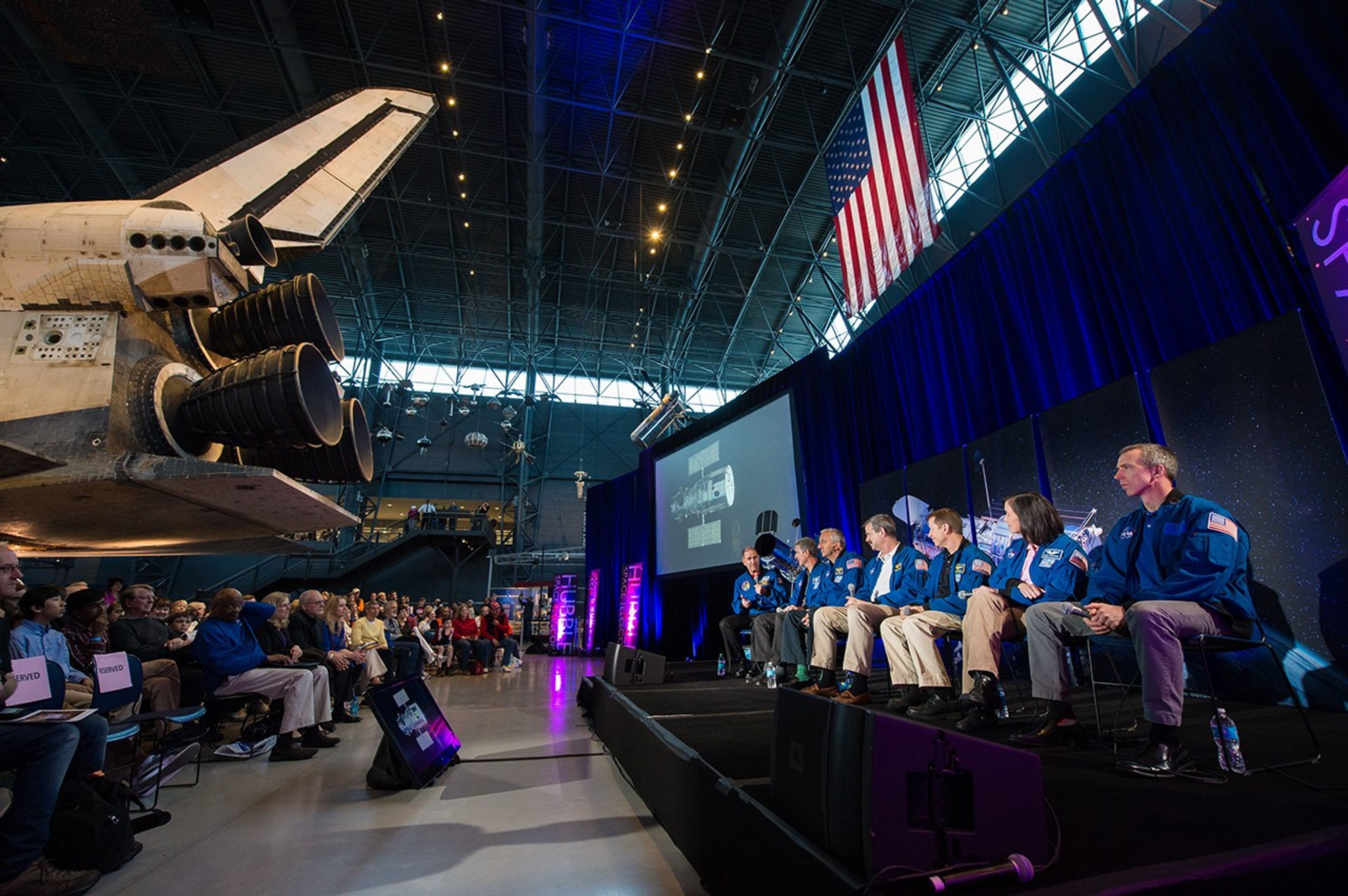 On the right half of the image is the astronaut panel onstage; John Grunsfeld, farthest to the left, speaks to the audience while facing his fellow astronauts, who look toward him. On the left side of the image, the audience listens attentively, sitting underneath the back of the Discovery Space Shuttle.