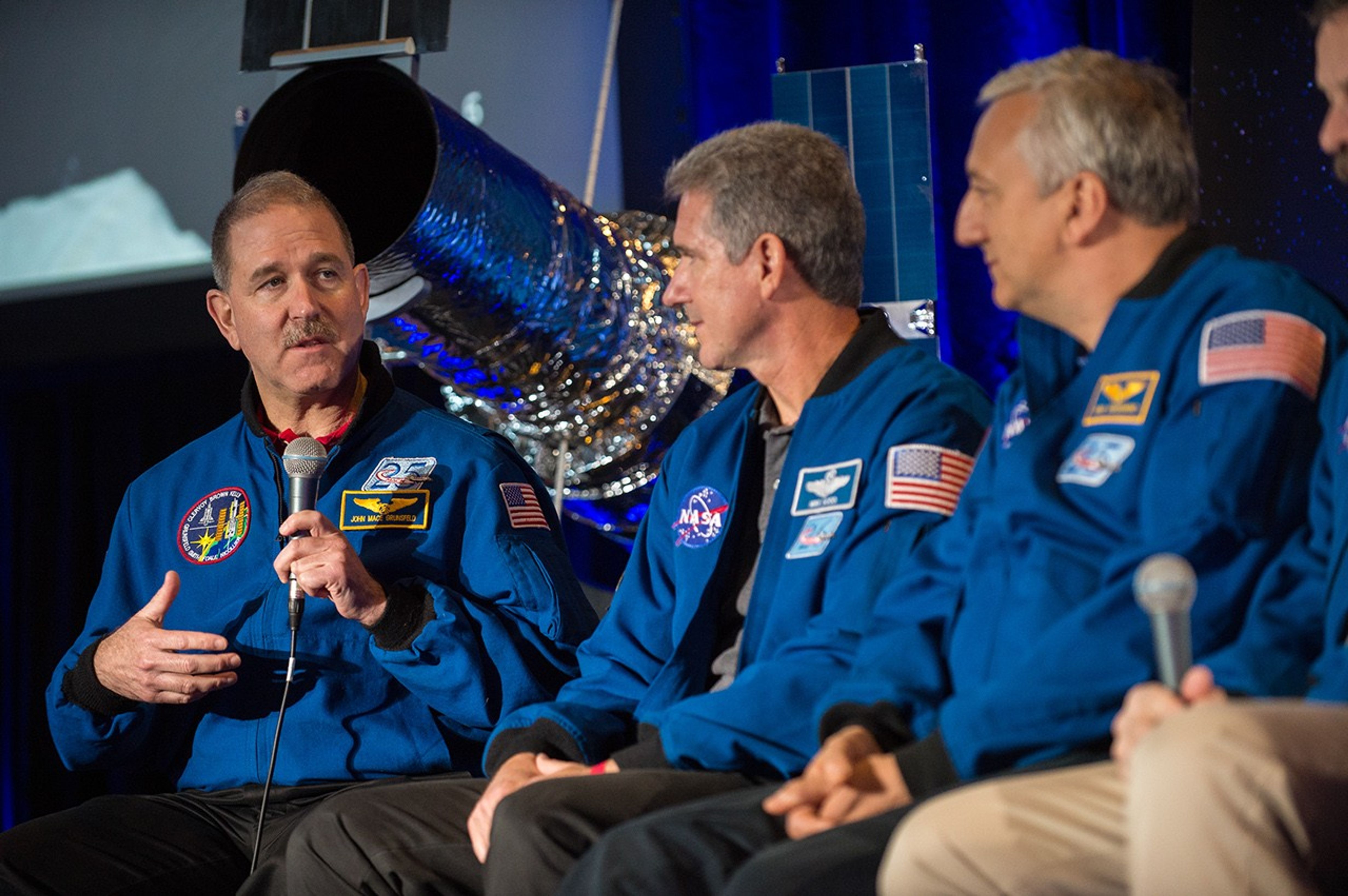 John Grunsfeld, left, addresses the audience, while his fellow astronauts look toward him. The Hubble model is visible in the background.