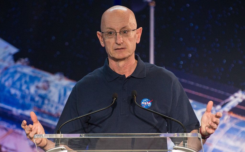 Eric Smith stands at the podium, addressing the crowd. Behind him in the background, part of a large image of the Hubble Space Telescope is visible.