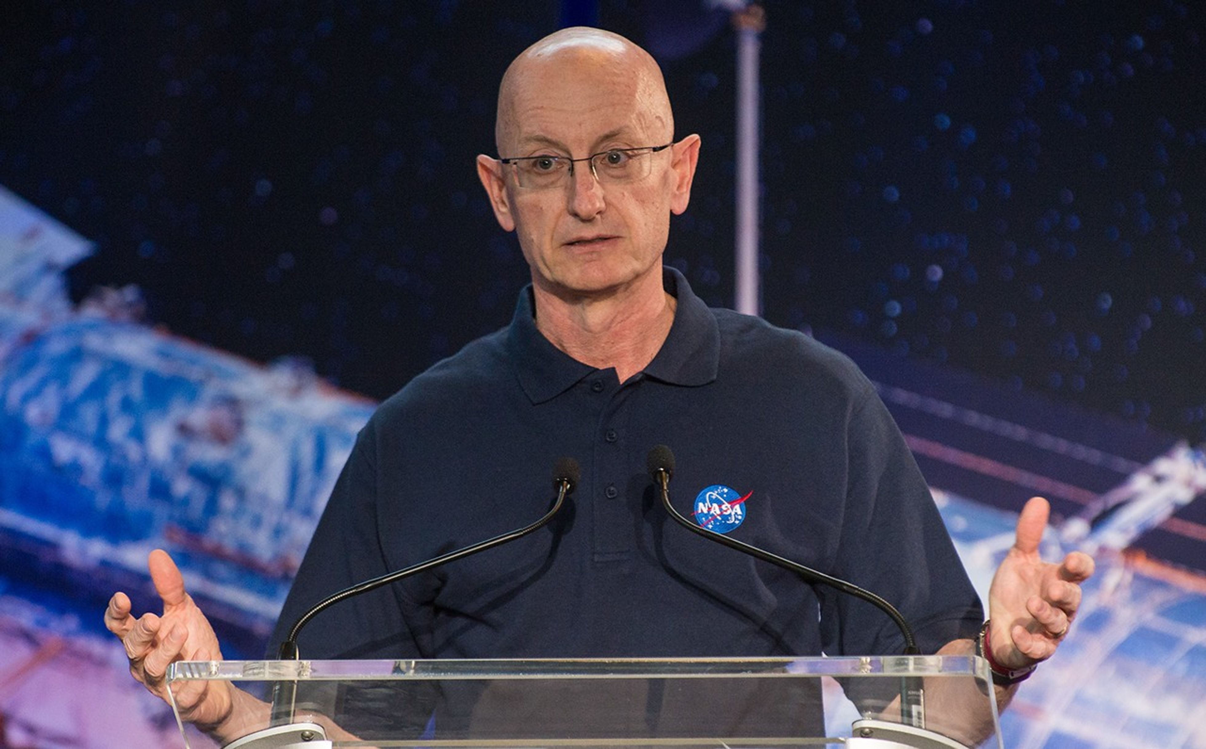 Eric Smith stands at the podium, addressing the crowd. Behind him in the background, part of a large image of the Hubble Space Telescope is visible.