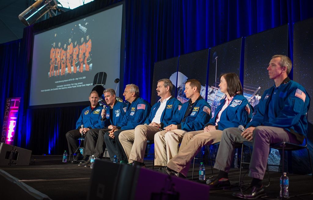 The astronauts on the panel sit onstage with a model and image of Hubble in the background, as Michael Good (second from the left) addresses the crowd. In the background on the left is the official NASA image of the HST Servicing Mission 4 astronauts