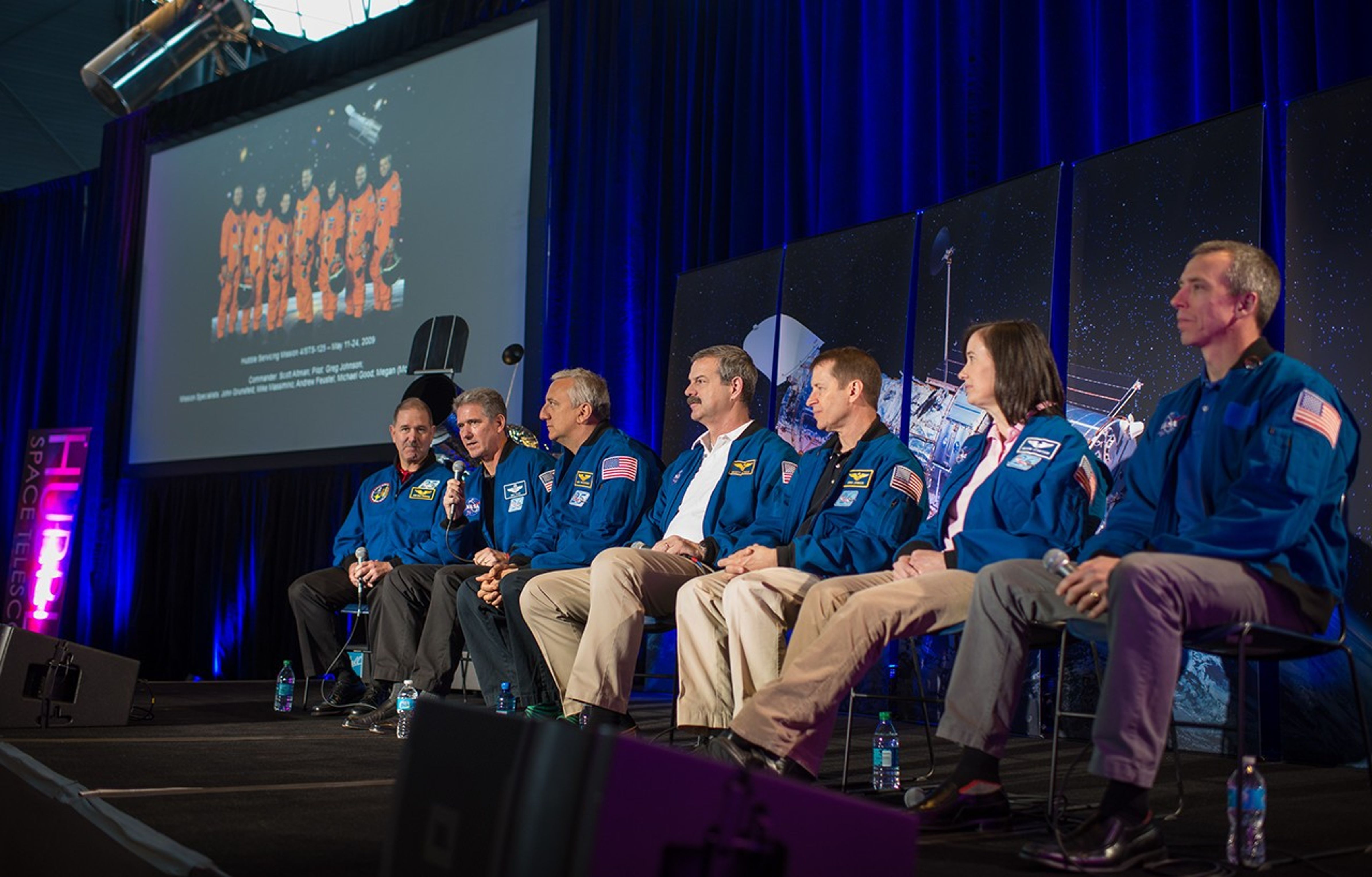 The astronauts on the panel sit onstage with a model and image of Hubble in the background, as Michael Good (second from the left) addresses the crowd. In the background on the left is the official NASA image of the HST Servicing Mission 4 astronauts