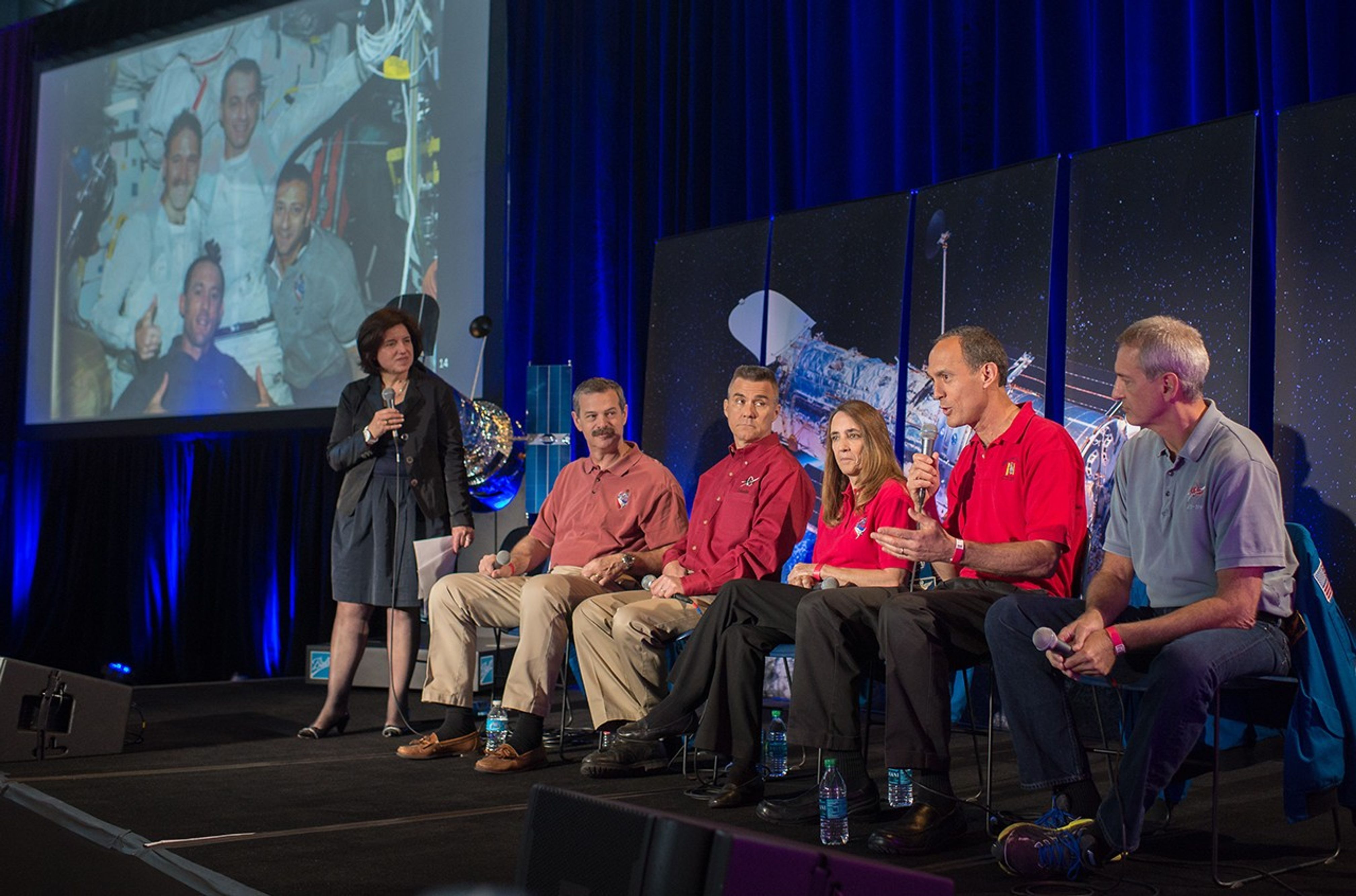 Five astronauts sit on the panel onstage, with Newman (second from the right) addressing the crowd while the other panelists and moderator Nota listen. On the left is a photo of four of the astronauts in the space shuttle on Servicing Mission 3B.