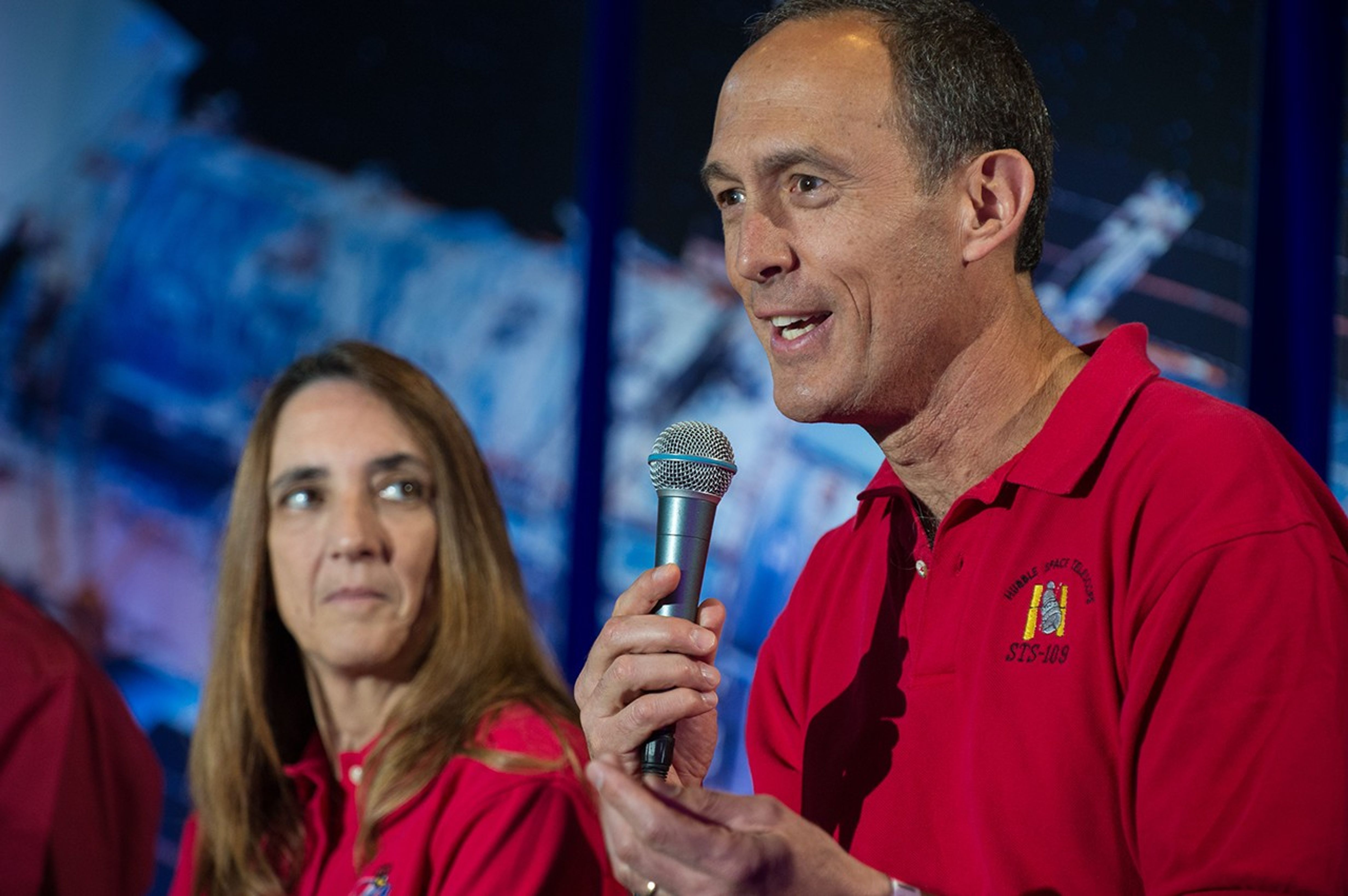 Jim Newman (right) addresses the audience, while in the background, fellow astronaut Nancy Currie (left) listens.