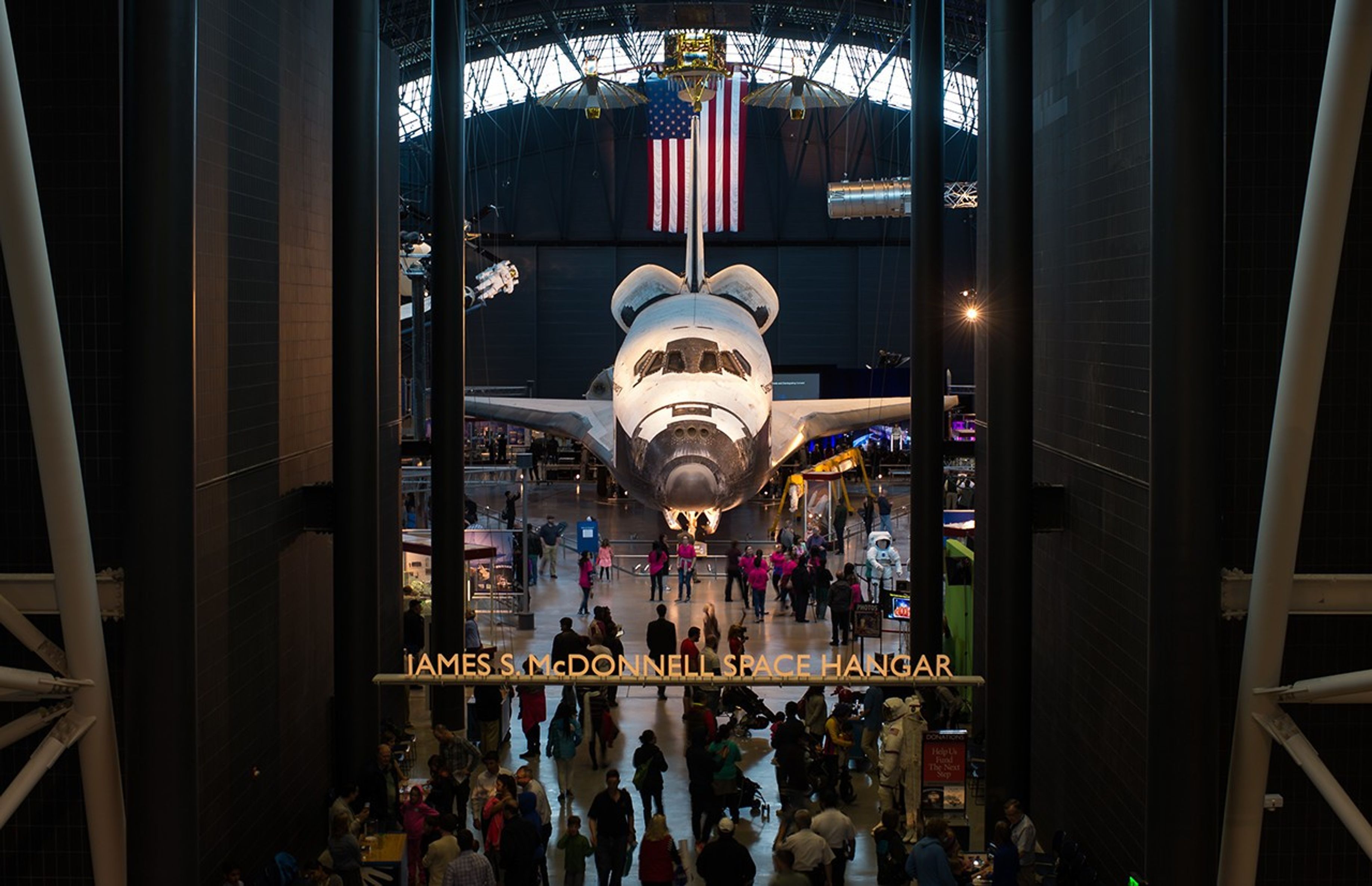 An image taken from above (the second floor) looking into the McDonnell Space Hangar. Many visitors are visible outside and inside the gallery. Visible within the gallery is the Discovery Space Shuttle and other exhibit displays. a US flag and models of several spacecraft (including Hubble) hang from the ceiling).