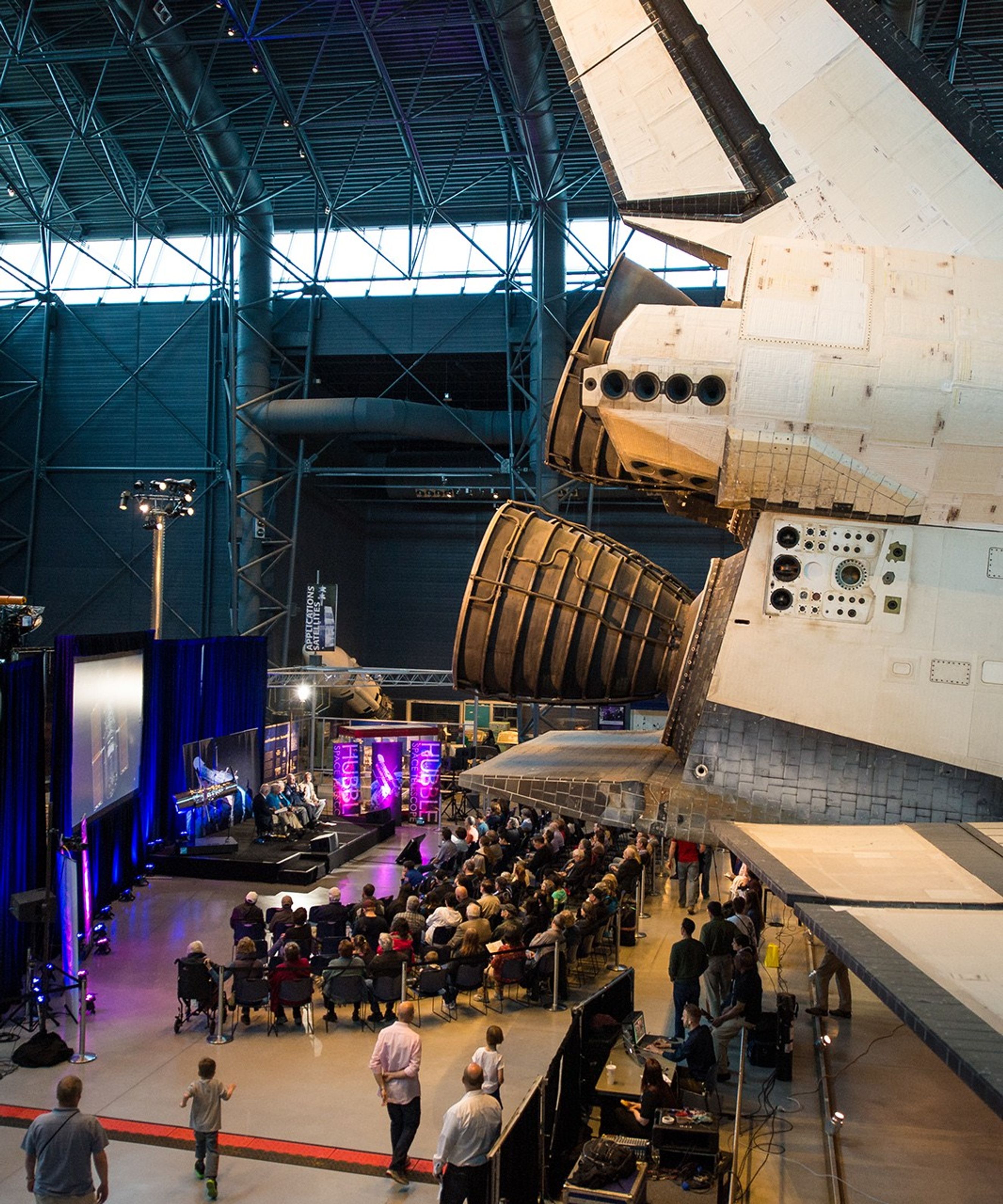A photo taken from above and from a distance. The right half of the picture is mostly taken up by the back of Space Shuttle Discovery in the foreground. On the left, the astronauts sit on stage for the panel, with a model of Hubble behind them to the left, a large audience listening, and a film crew ensuring technology works correctly.