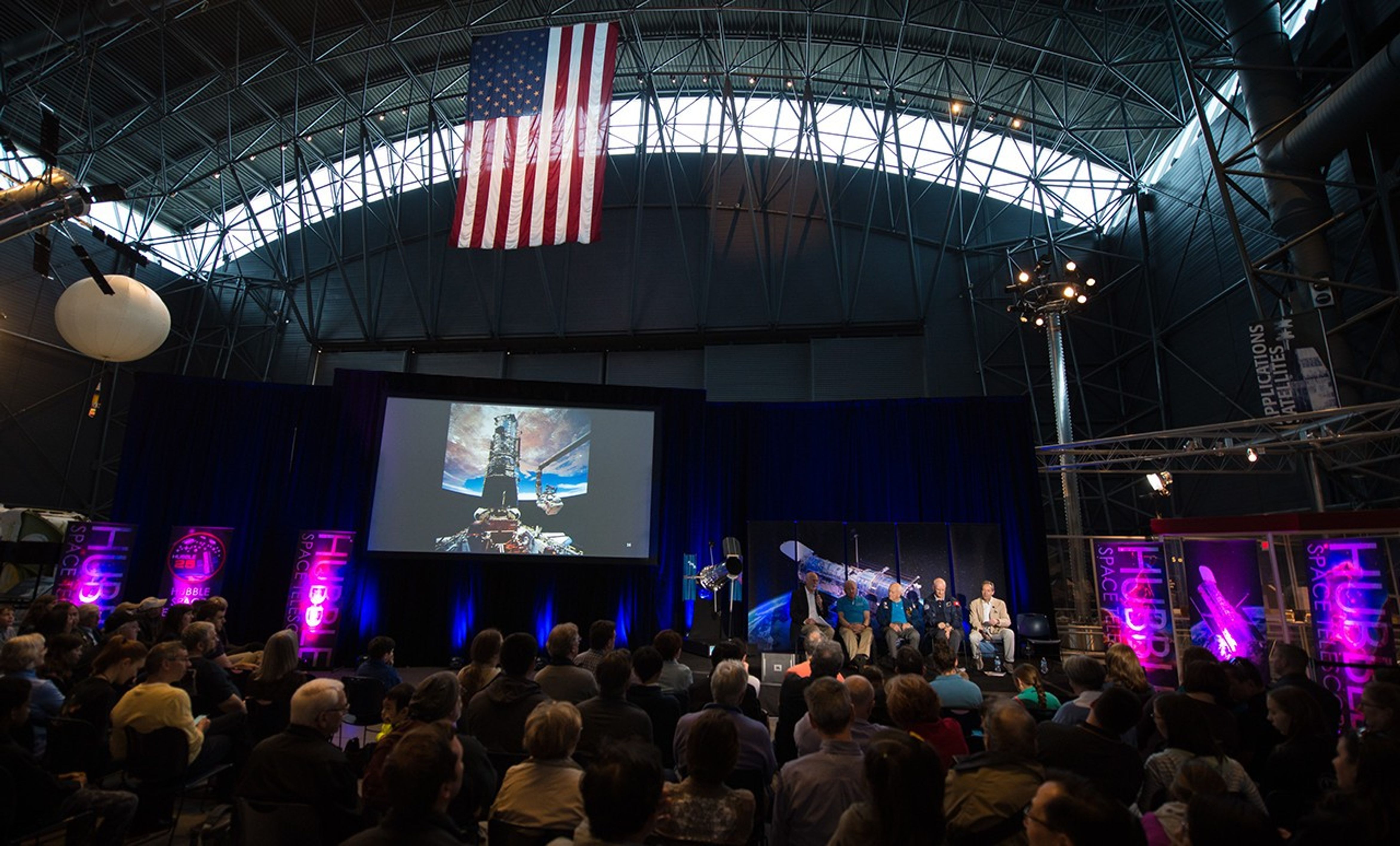 An image of the astronaut panel onstage taken from farther back in the audience. The audience members listen attentively. To the left of the astronauts is the Hubble model, and to the left of that, a large screen displaying images from Hubble servicing missions.