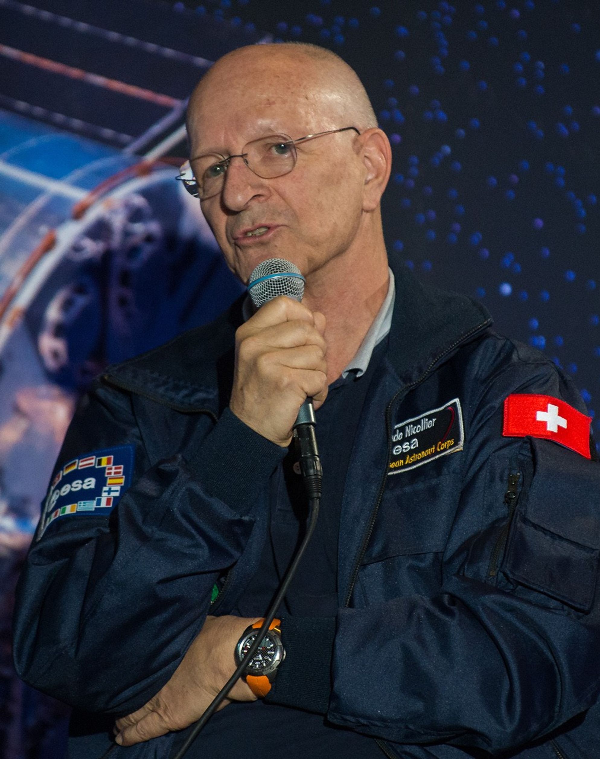 Astronaut Claude Nicollier holds a microphone and addresses the audience.