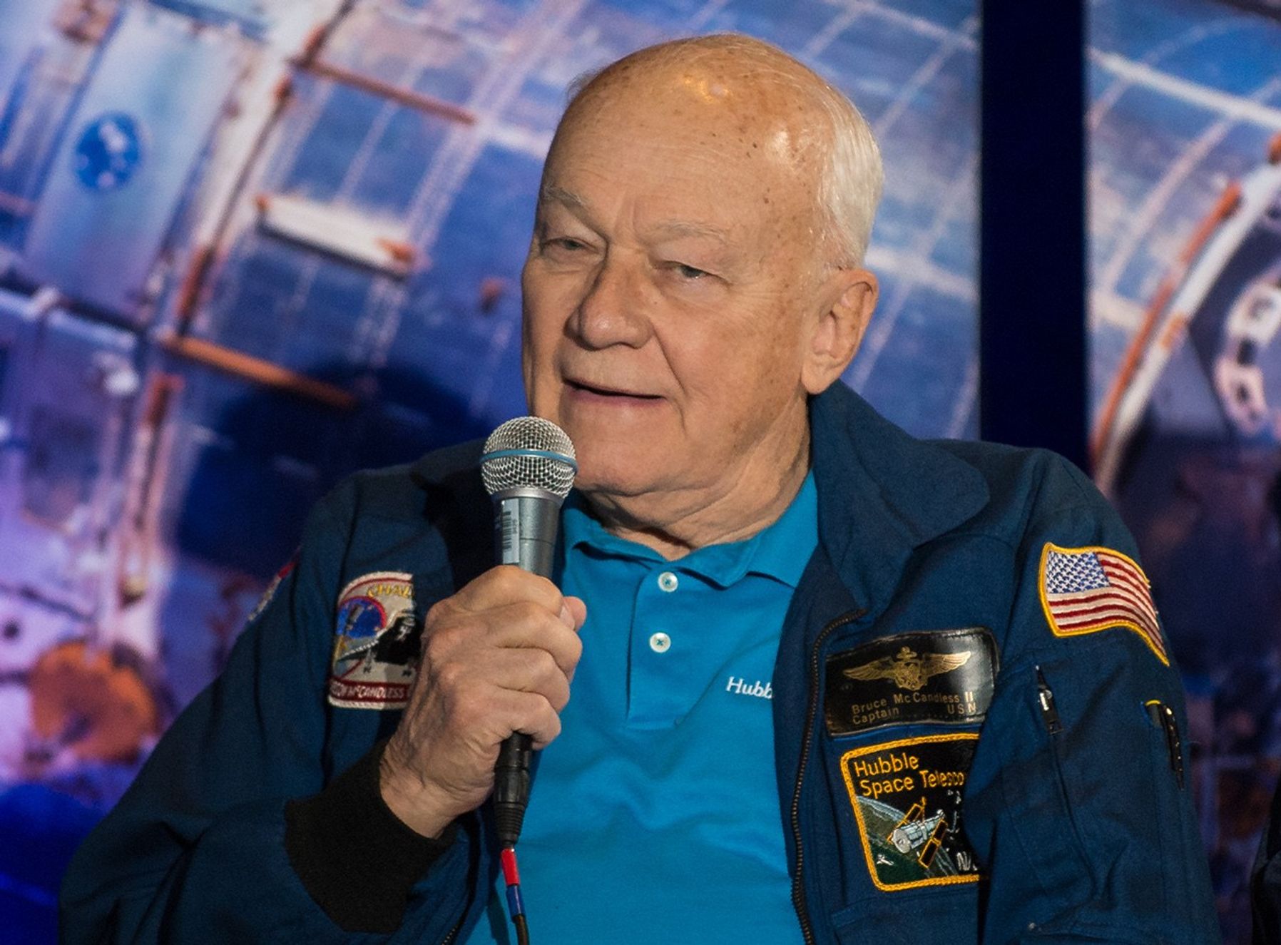 Bruce McCandless holds a microphone and addresses the audience.