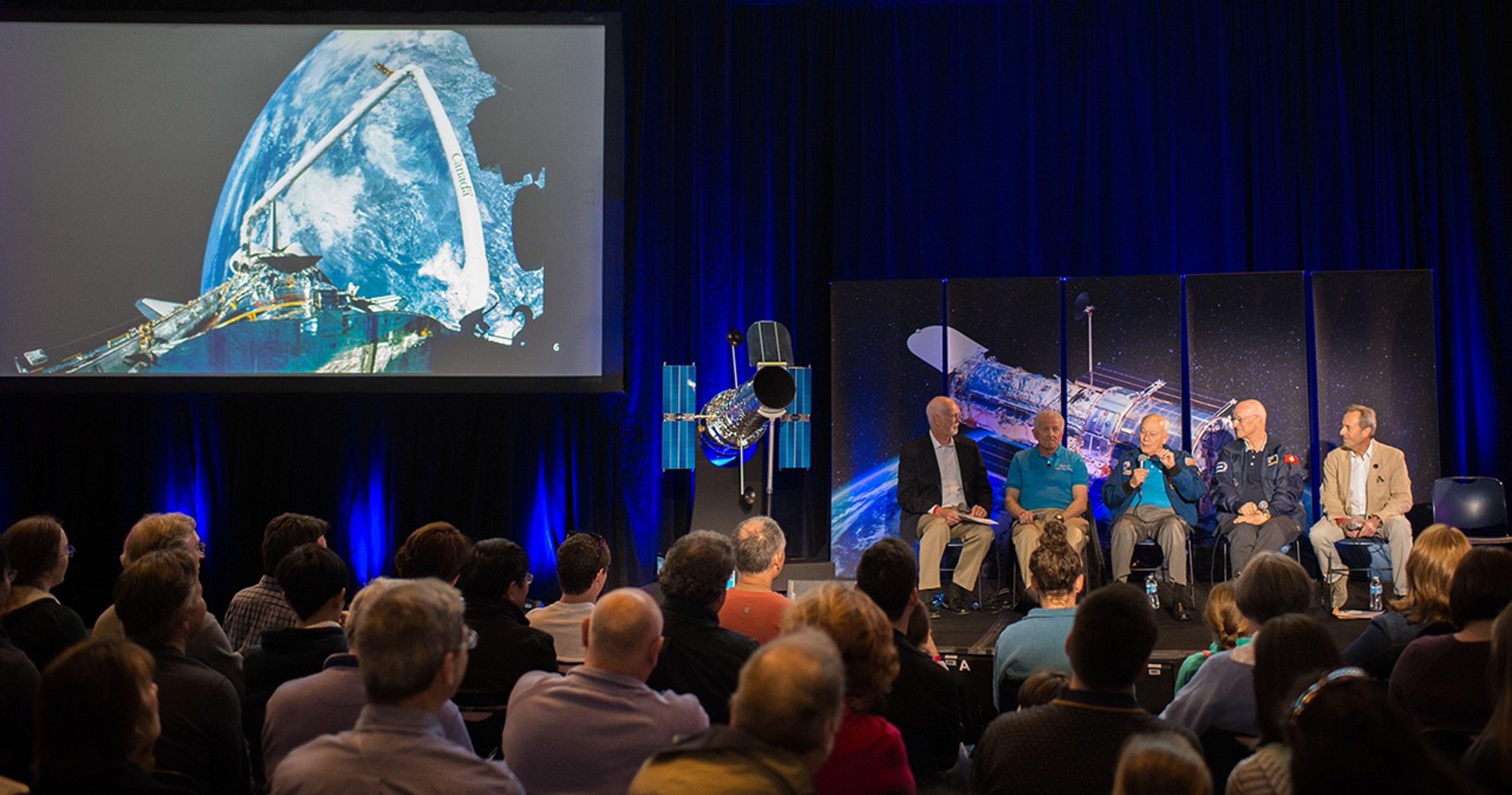 An image of the astronaut panel onstage taken from a few rows back in the audience. The audience members listen attentively. To the left of the astronauts is the Hubble model, and to the left of that, a large screen displaying images from Hubble servicing missions.