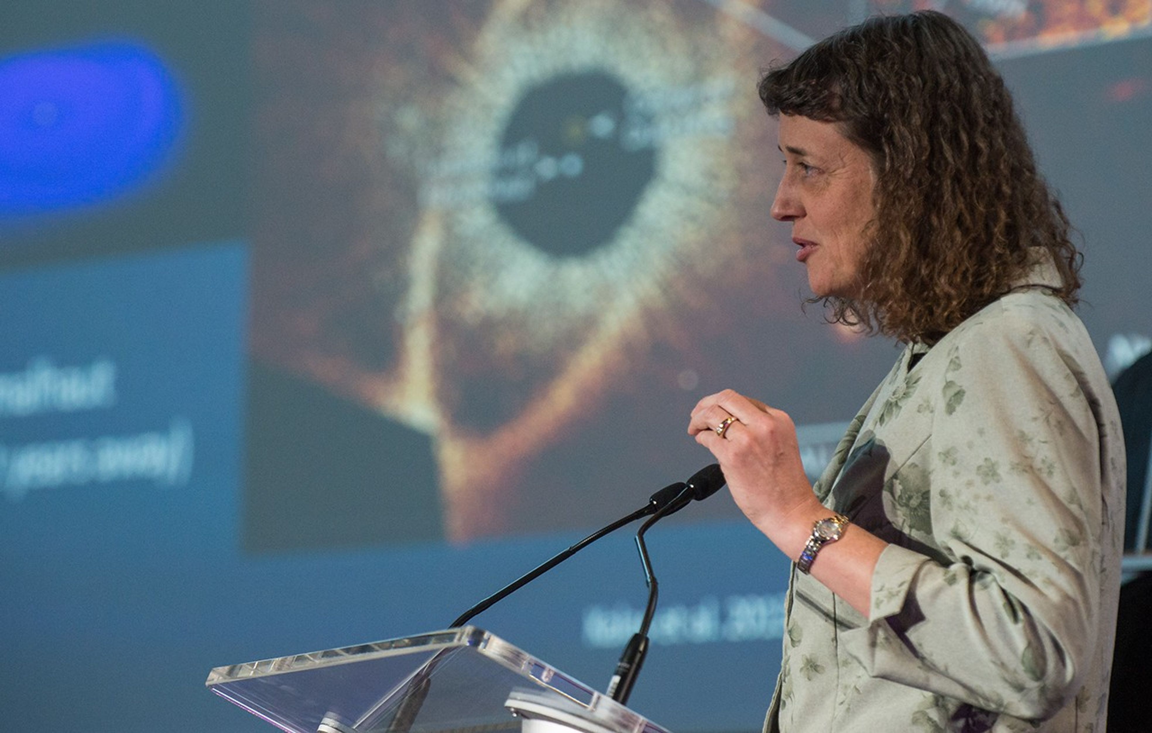 Dr. Jennifer Wiseman stands at the podium, speaking to the audience. In the background is a presentation on the big screen, with a Hubble astronomical image visible.