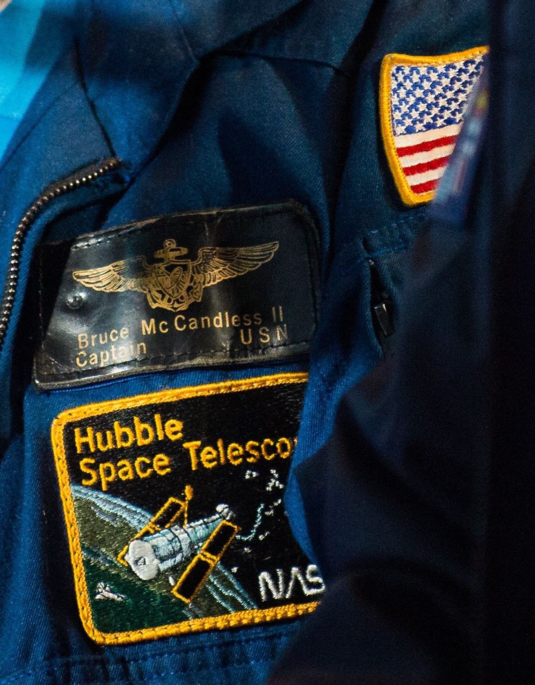 A photo of the mission patches on Bruce McCandless's flight jacket. The top patch has is name and rank on it. The bottom one is a Hubble Space Telescope patch showing Hubble over the Earth. In the upper-right corner, part of the American flag patch on McCandless's left arm is visible.
