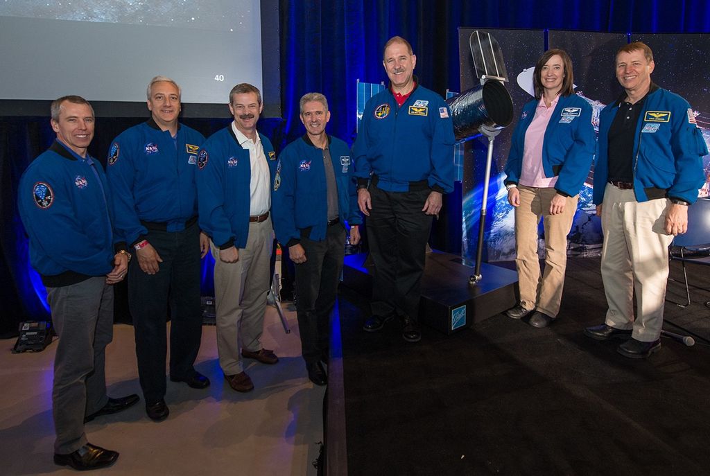 Seven astronauts, six men and one woman, from the 5th and final servicing mission (SM4) stand in a row; the three on the right are on the left side of the stage, while the four on the left are standing next to the stage. Behind them is the model of the Hubble Space Telescope, along with a banner image of it.