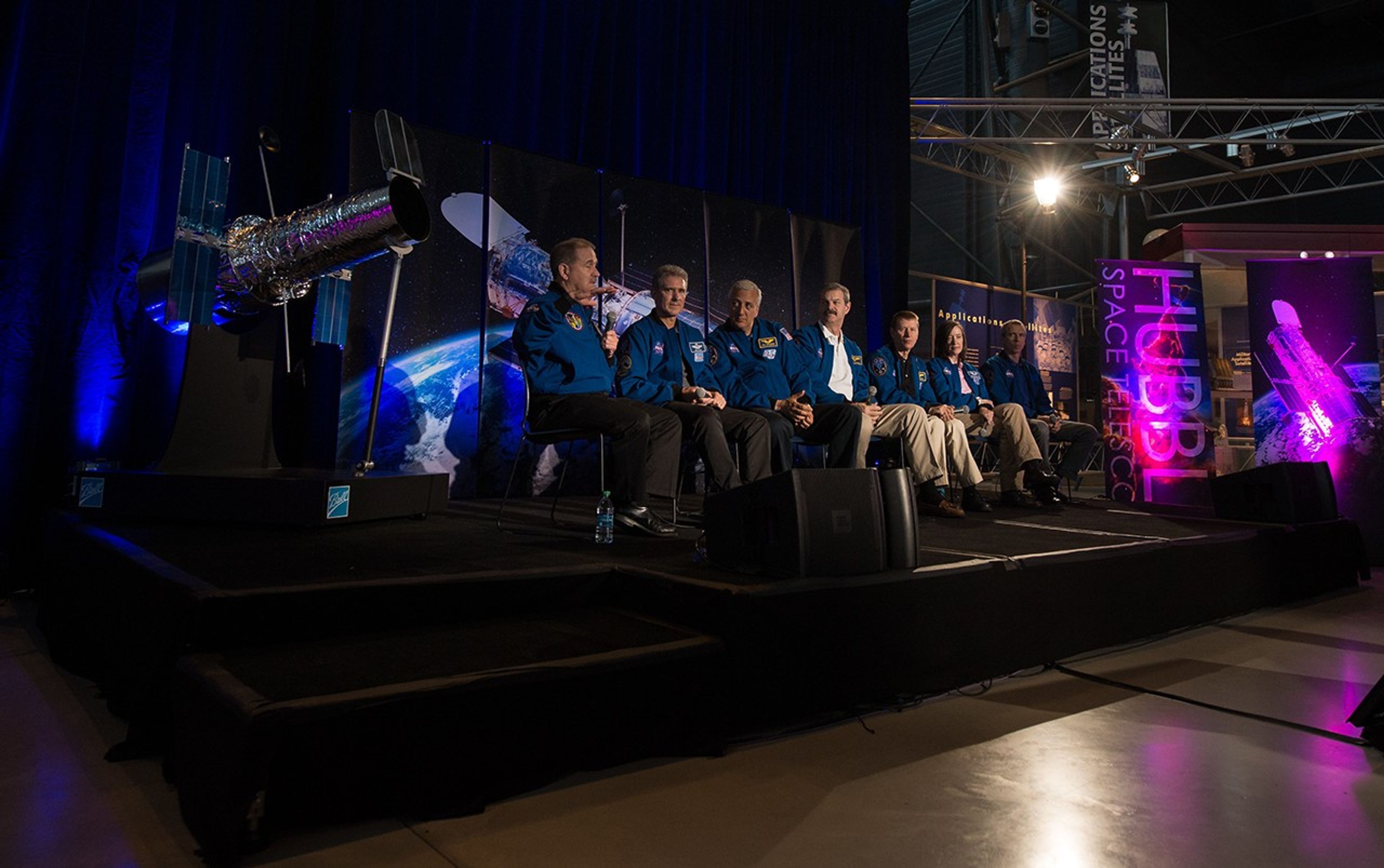 Seven astronauts sit in chairs on the stage, with a Hubble model on the left, an image of Hubble over the Earth behind them in the background, and other banners on the right. John Grunsfeld, the astronaut farthest to the left, speaks to the crowd, while the other astronauts listen.
