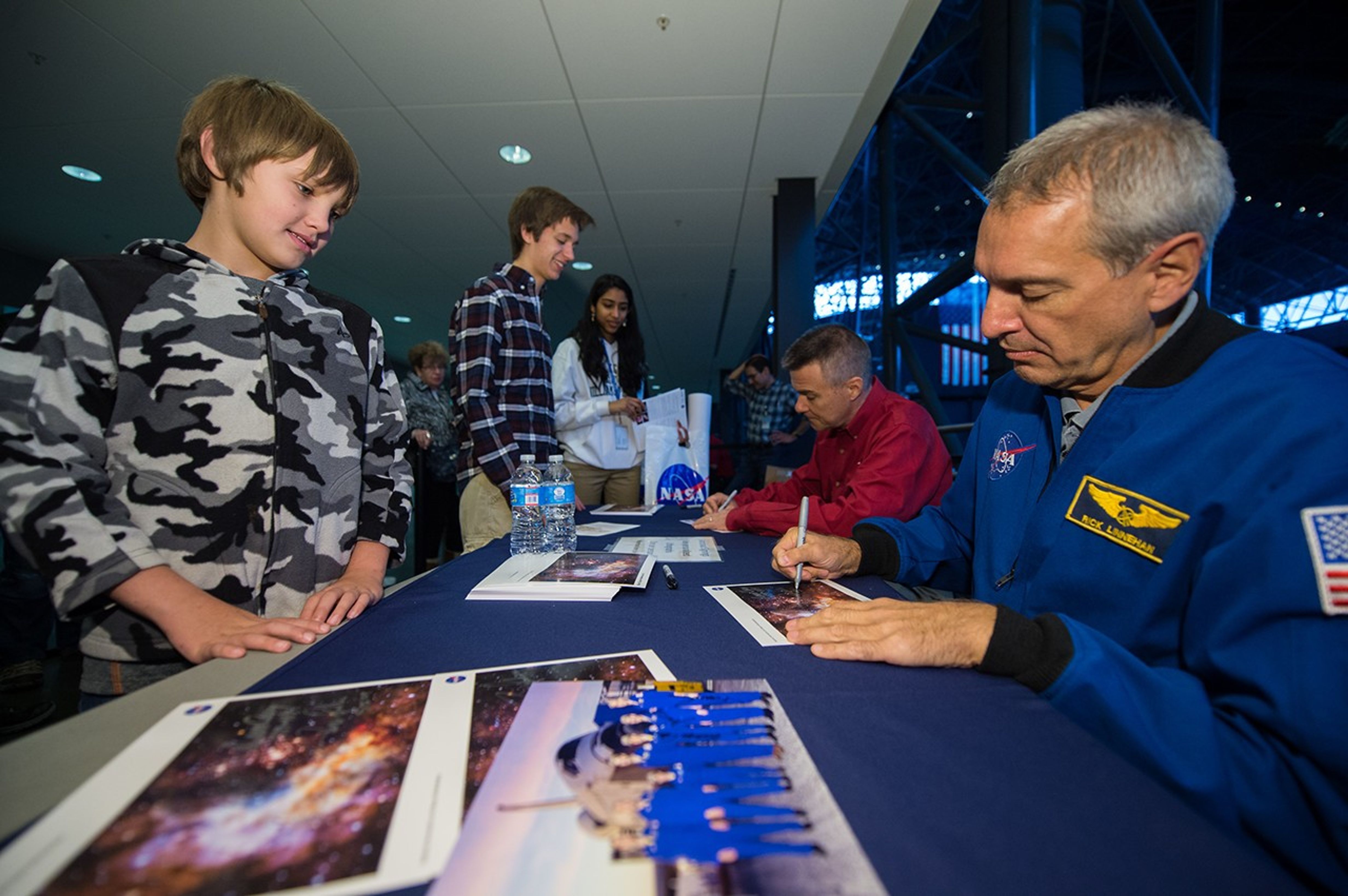 Astronauts Richard Linnehan and Dane Carey and  sit on the right-hand side of the table, signing lithographs and photos for visitors. On the left hand side are three young people receiving the signed pictures.