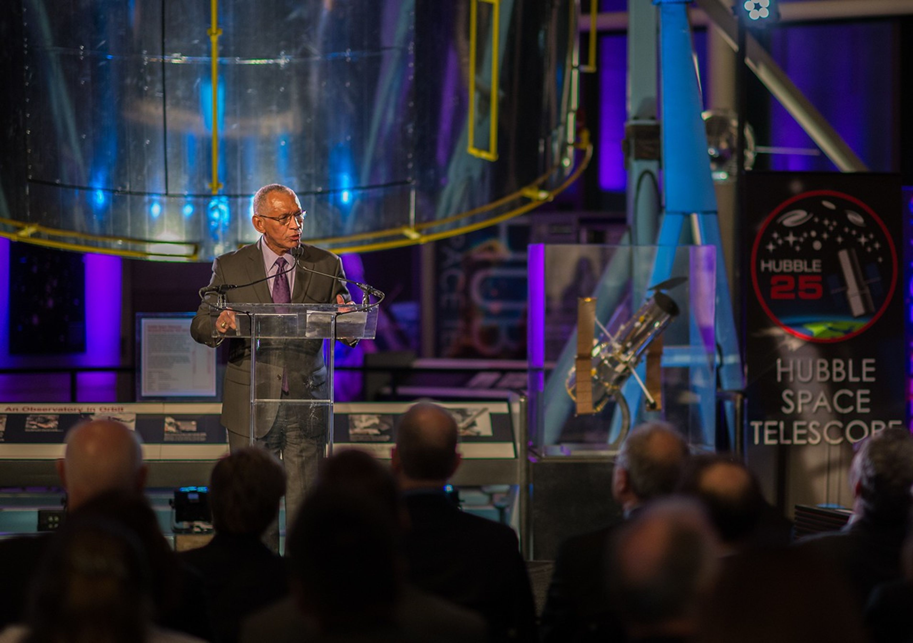 Charlie Bolden stands at the podium speaking to an attentive audience, with the Hubble Structural Dynamic Test Vehicle in the background, and a model of Hubble and 25th anniversary poster on the right.
