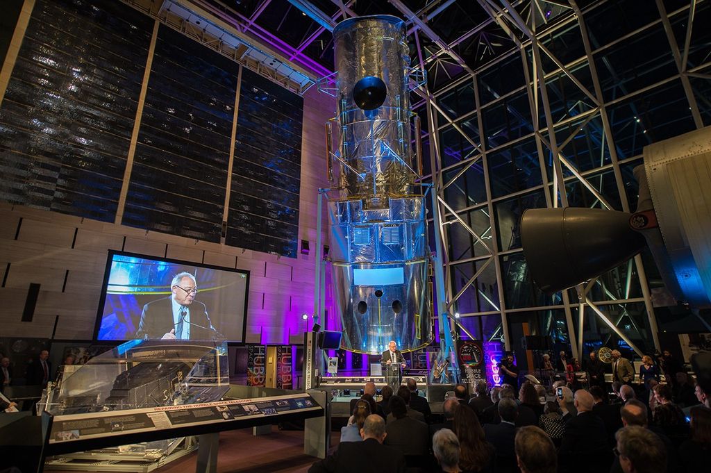 As Dordain addresses the audience, the Hubble Test Vehicle and clear glass wall and ceiling of the Air and Space Museum take up the background, along with other museum displays. The audience can be seen listening in the foreground.