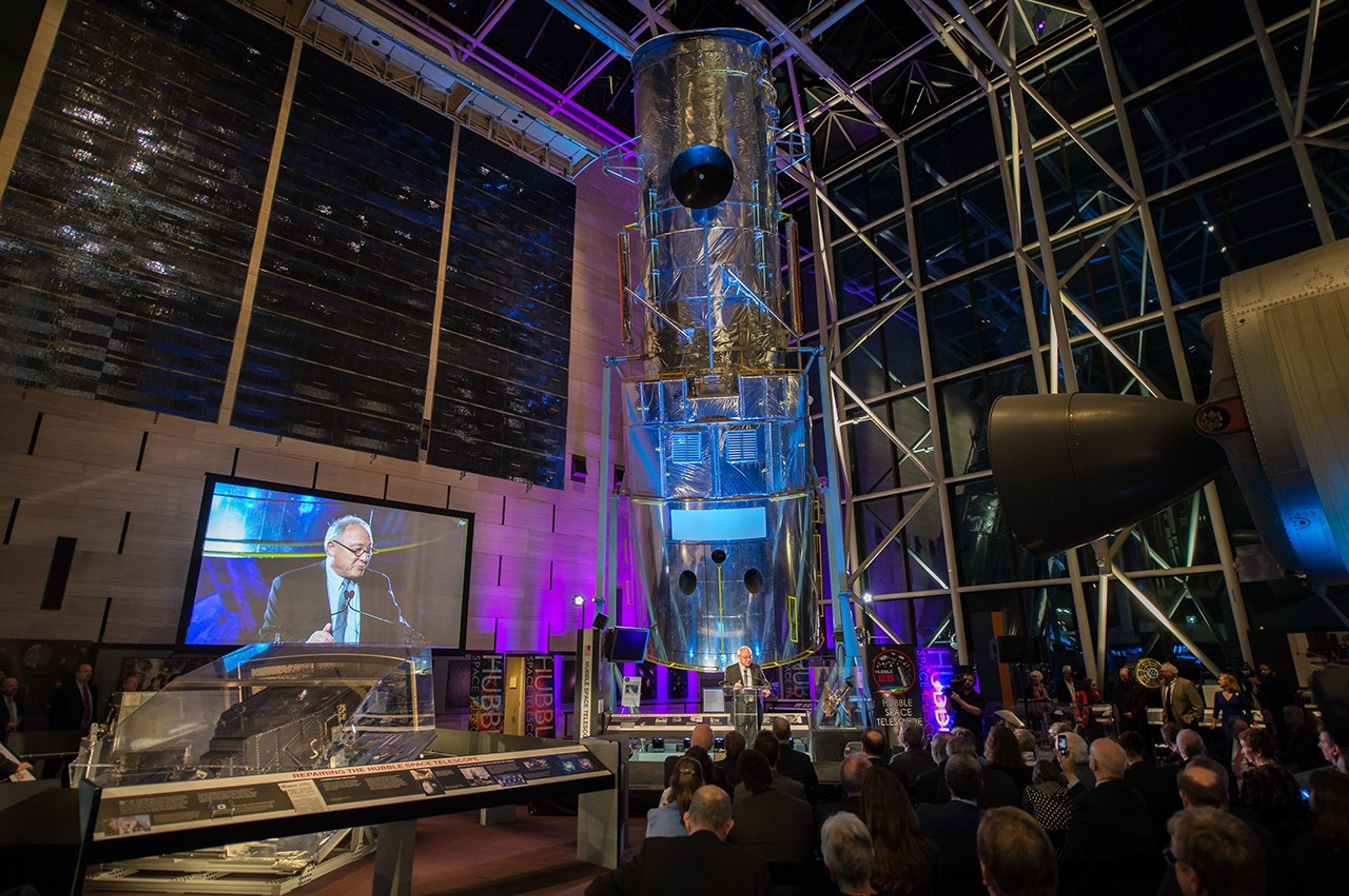 As Dordain addresses the audience, the Hubble Test Vehicle and clear glass wall and ceiling of the Air and Space Museum take up the background, along with other museum displays. The audience can be seen listening in the foreground.