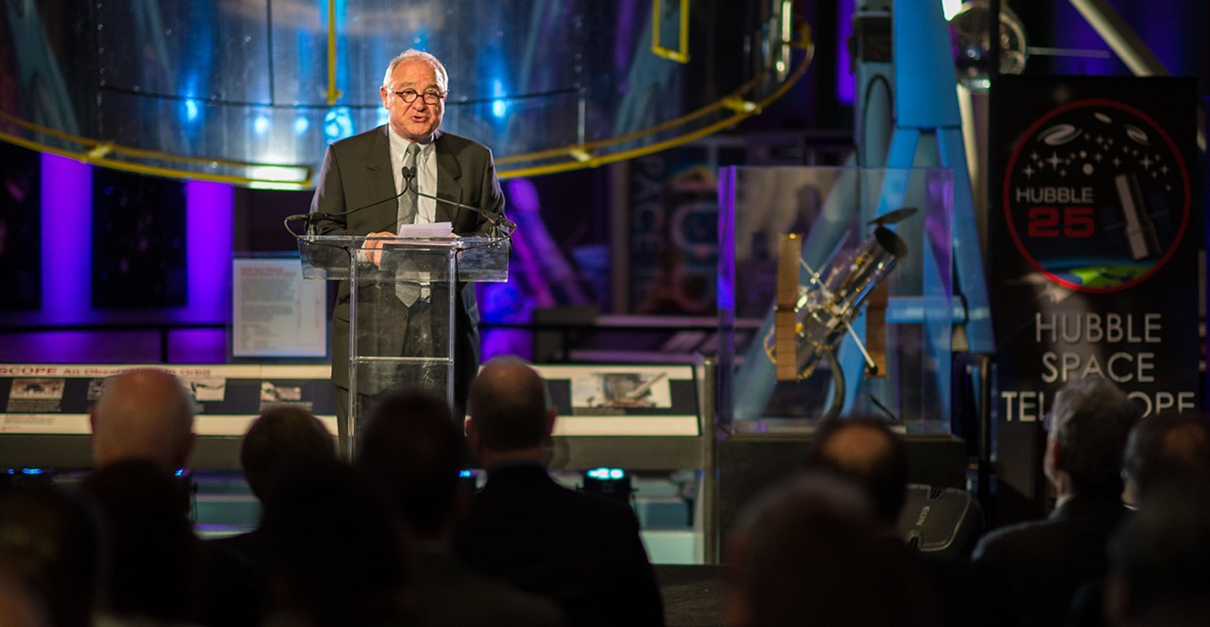 Jean-Jacques Dordain stands at the podium speaking to an attentive audience, with the Hubble Structural Dynamic Test Vehicle in the background, and a model of Hubble and 25th anniversary poster on the right.