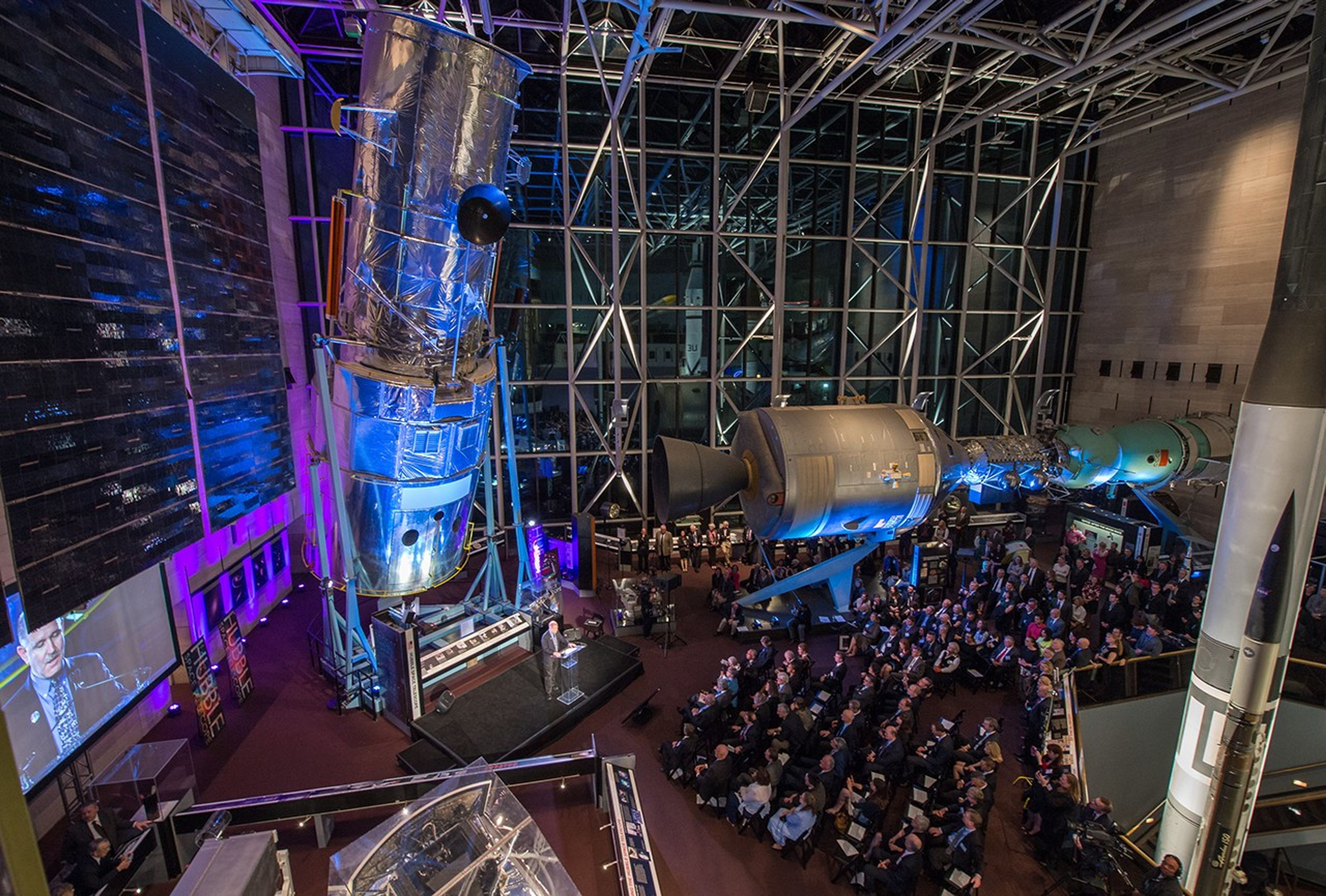 A wide view of the Hubble Test Vehicle and other displays in the interior of the Air and Space Museum, with a large audience listening to John Grunsfeld speak.