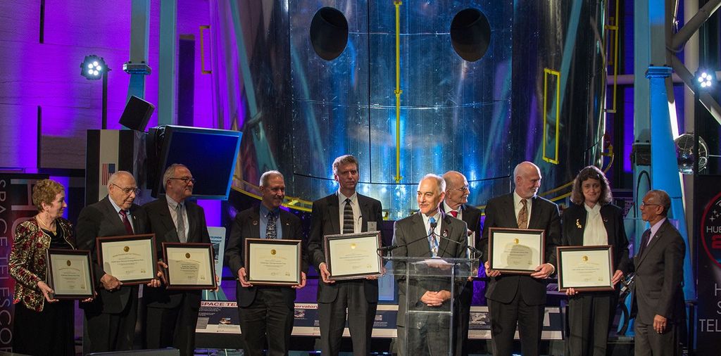 The eight award recipients stand in a row on the stage, with Charlie Borden standing to the right of the last winner, Dr. Wiseman. Another man stands in front of them at the podium.