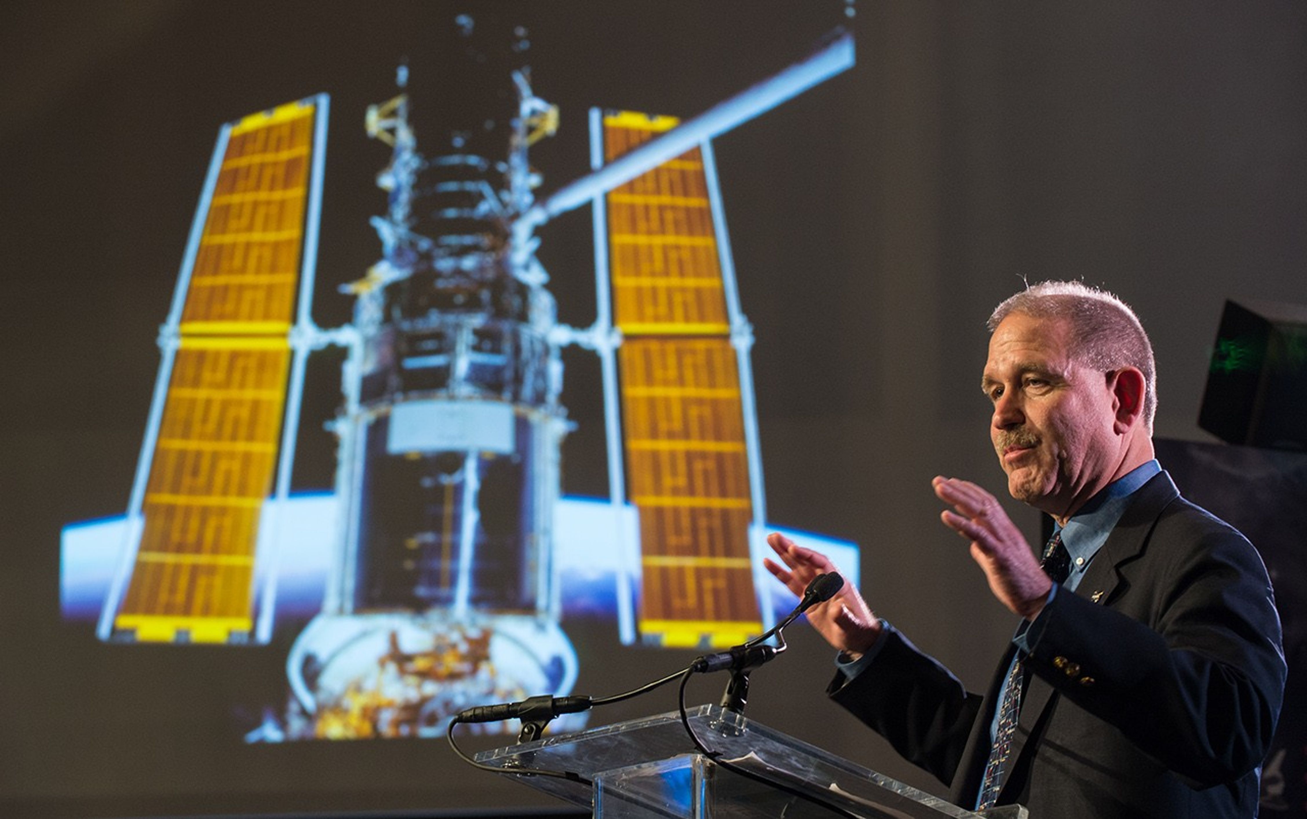 John Grunsfeld stands at the podium, addressing the audience, with an image of Hubble onscreen in the background.