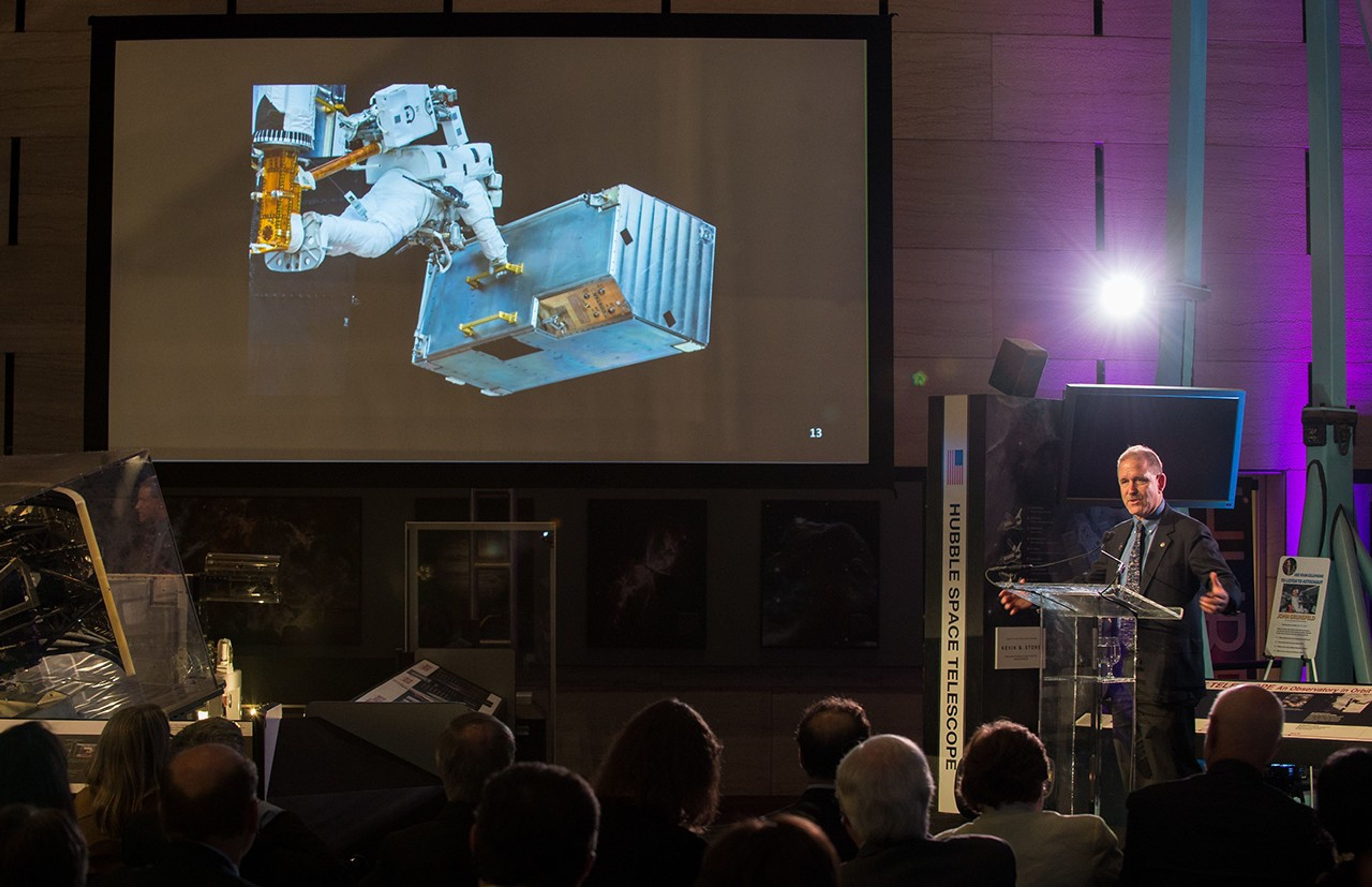 John Grunsfeld stands at the podium, addressing the listening audience. Above and to the left of him, the screen shows an image of a astronaut on a Hubble servicing mission. Other museum displays can be seen on the left and right.