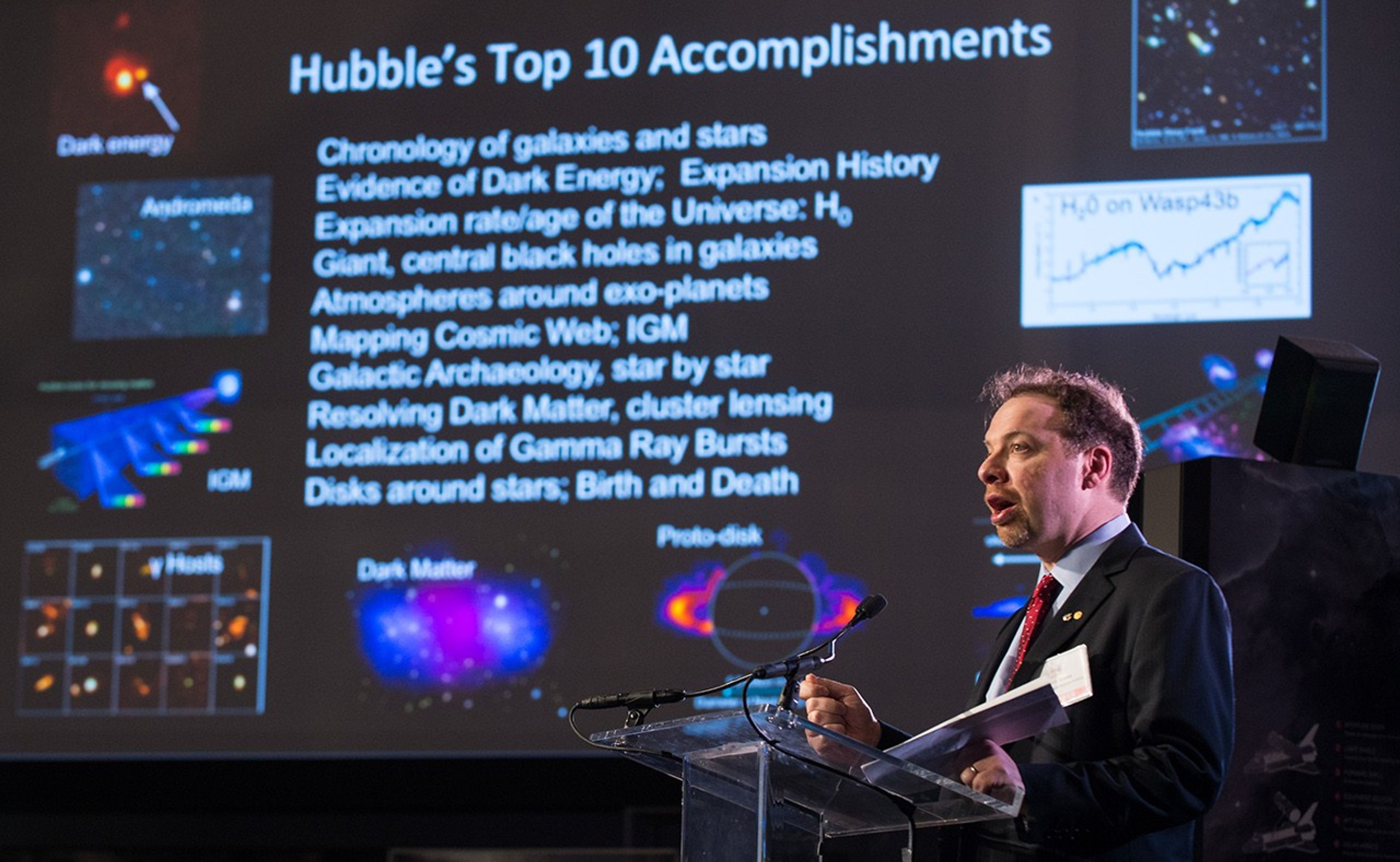 Adam Riess stands at the podium, addressing the audience. In the background, a slide about Hubble's Top 10 accomplishments can be seen on the screen.