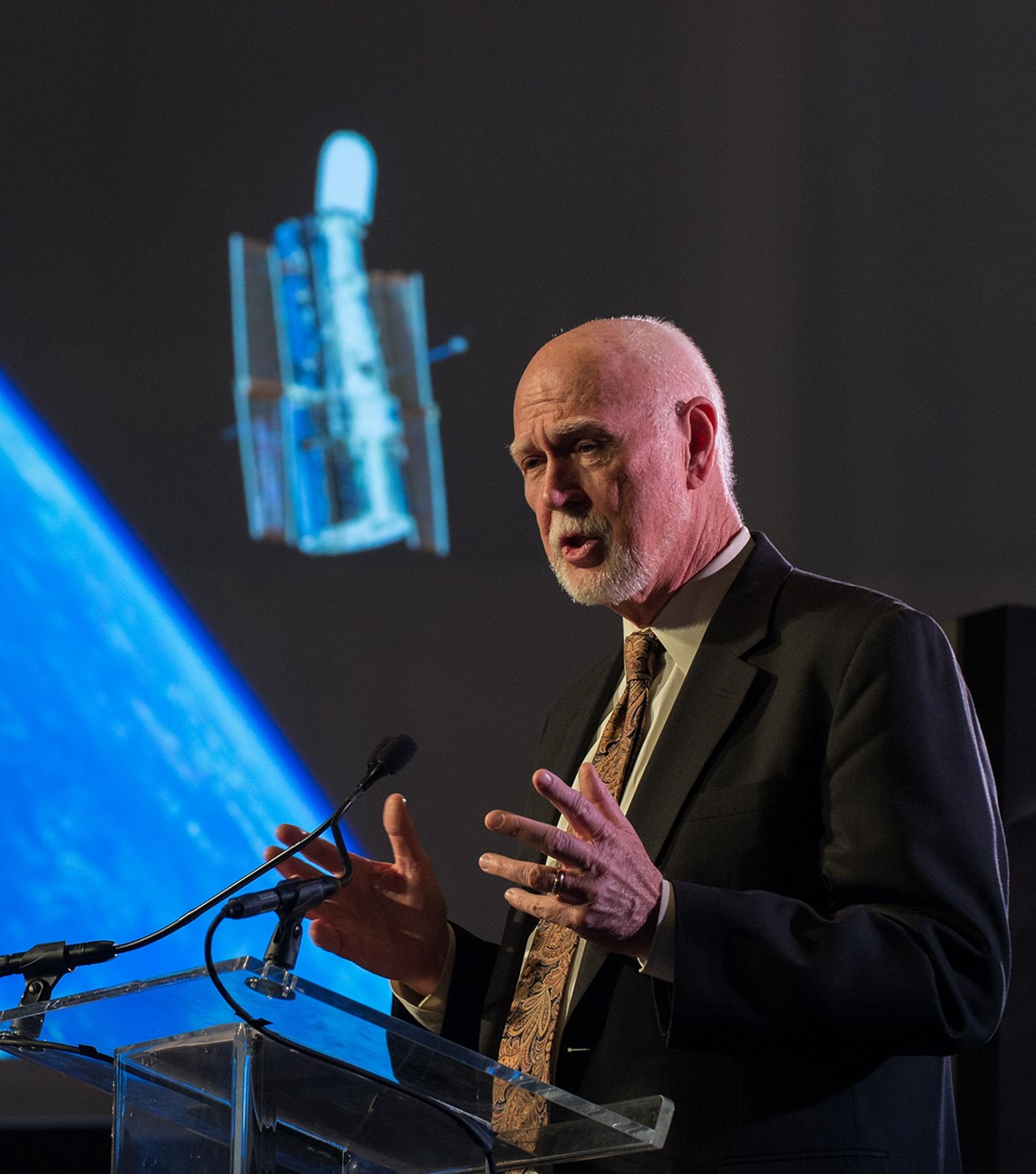 Jim Crocker stands at the podium, addressing the audience. In the background, a photo of Hubble over the Earth can be seen on the screen.
