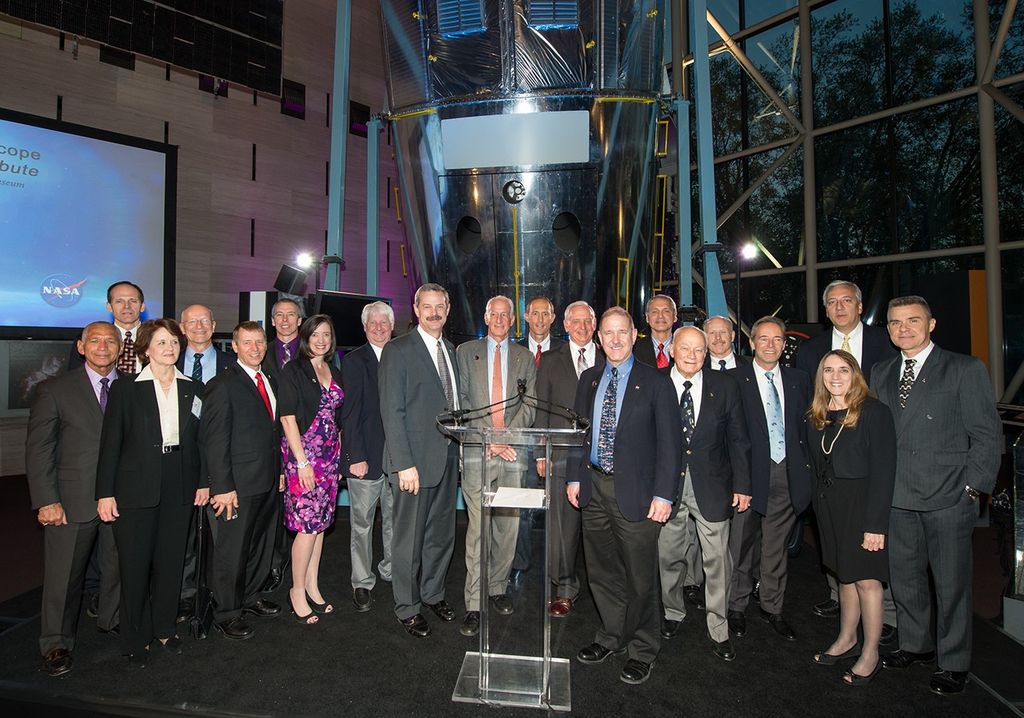 A group of 20 men and women stand in front of the Hubble test vehicle behind a clear podium, with a slide show prepared on a screen on the left.