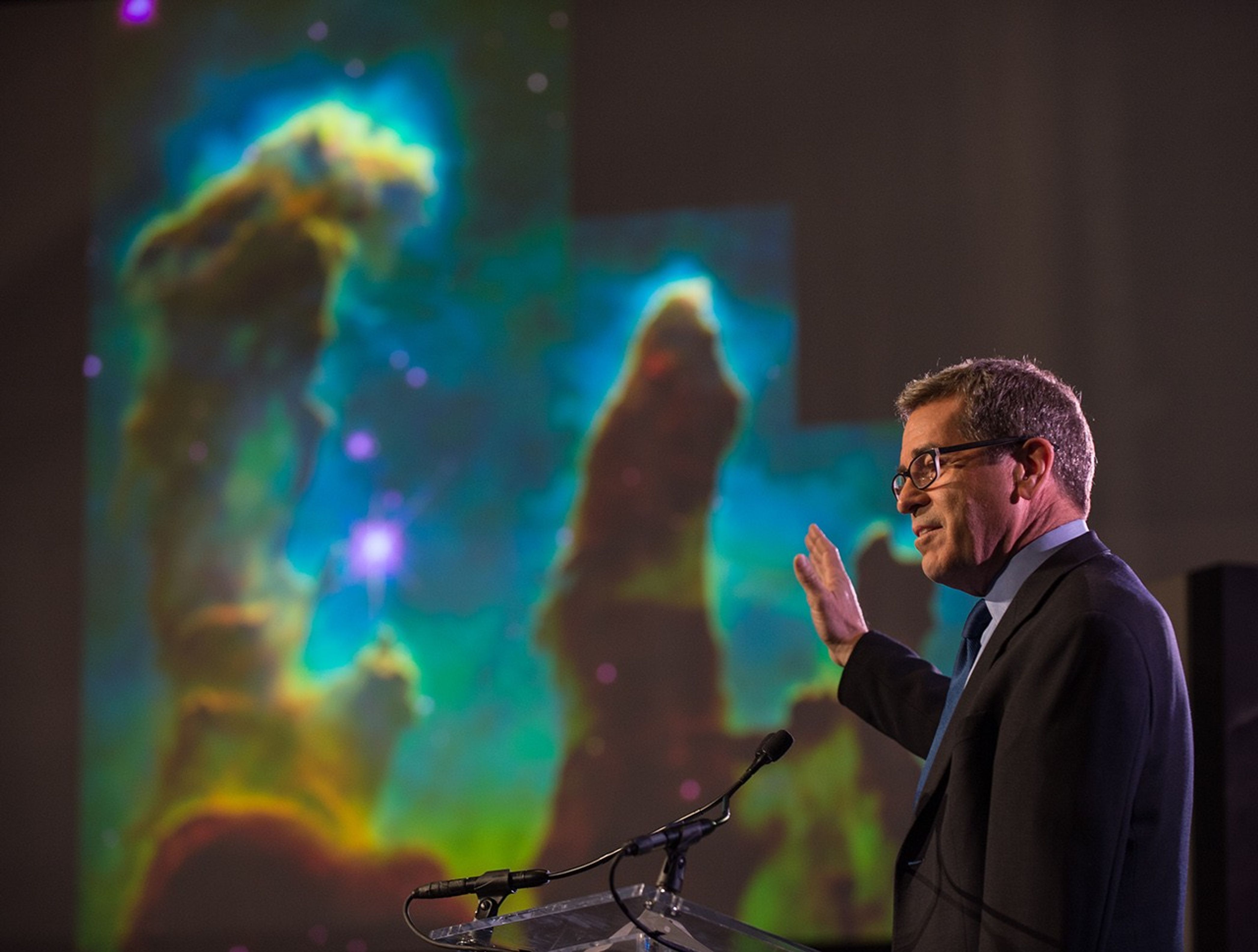 Miles O'Brien stands at the podium, with a Hubble image of the Pillars of Creation in the background.