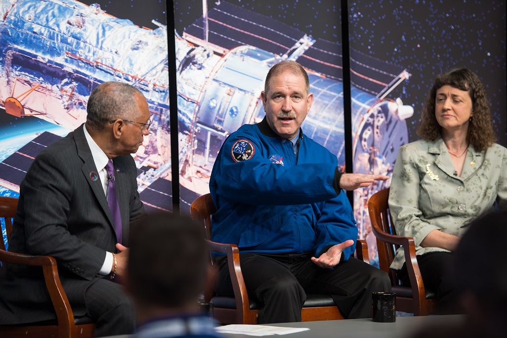John Grunsfeld (center) looks at the camera and addresses the audience from his panelist's chair onstage, with Charlie Bolden on the left and Jennifer Wiseman on the right. A large image of Hubble over the Earth forms the backdrop behind them.