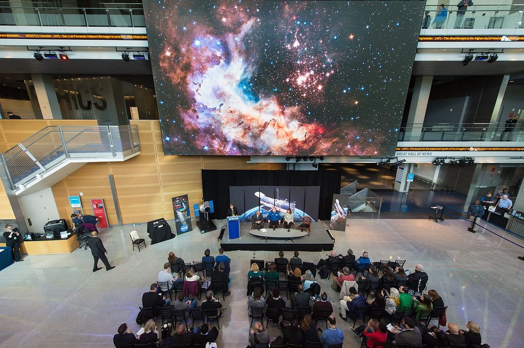 A wide shot taken from one floor above. The anniversary image of Westerlund 2 on the giant screen takes up the upper half of the picture. In the lower half, Dr. Flanagan speaks at the podium onstage while Bolden, Grunsfeld, and Dr. Wiseman sit in their panelist seats. An audience of around 50 people sits in chairs facing the stage.