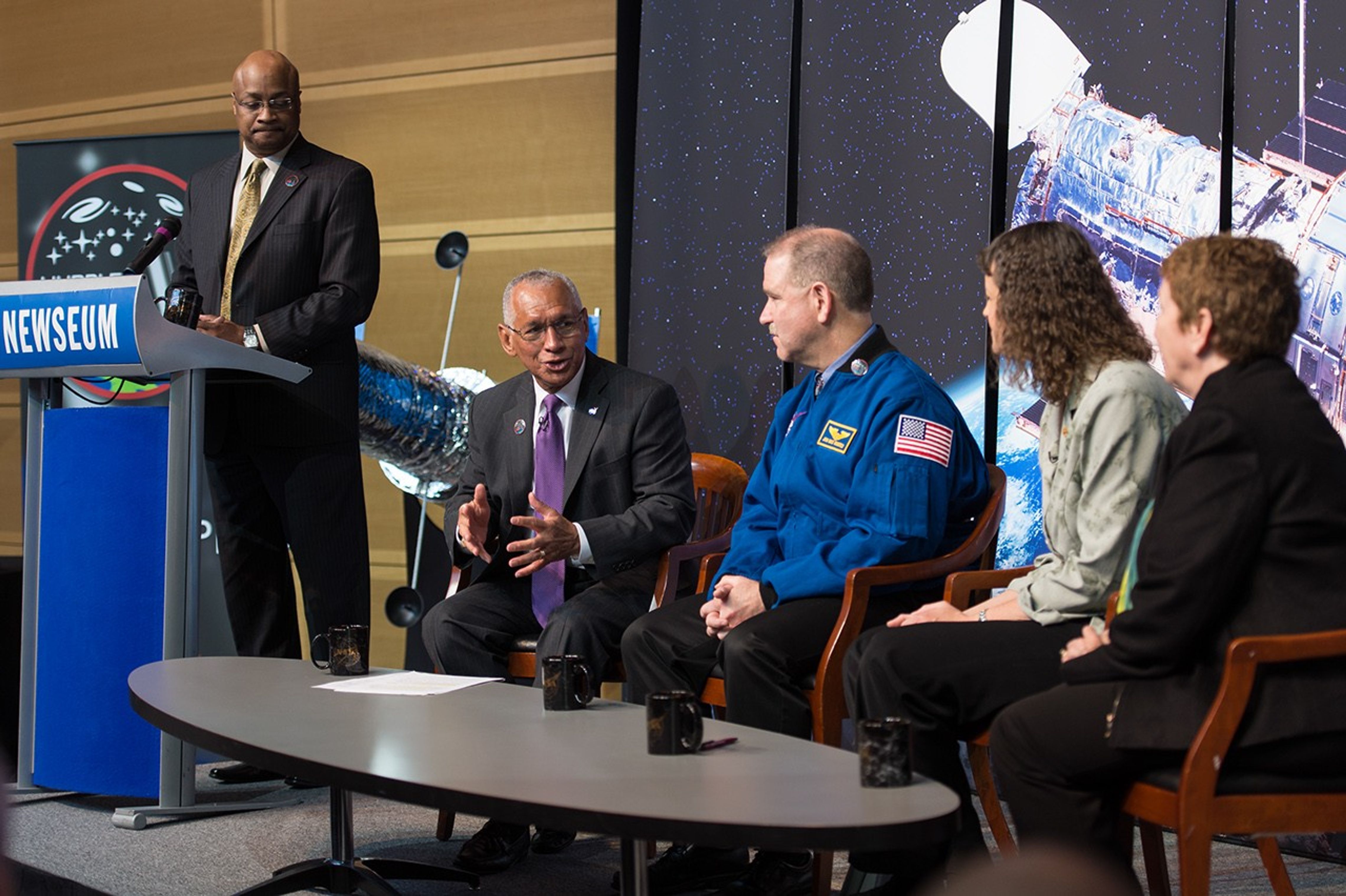 At the table of four panelists, Charlie Bolden (left) speaks to fellow three panelists John Grunsfeld, Jennifer Wiseman, and Kathryn Flanagan. Another unnamed gentleman stands on the far left at the podium.