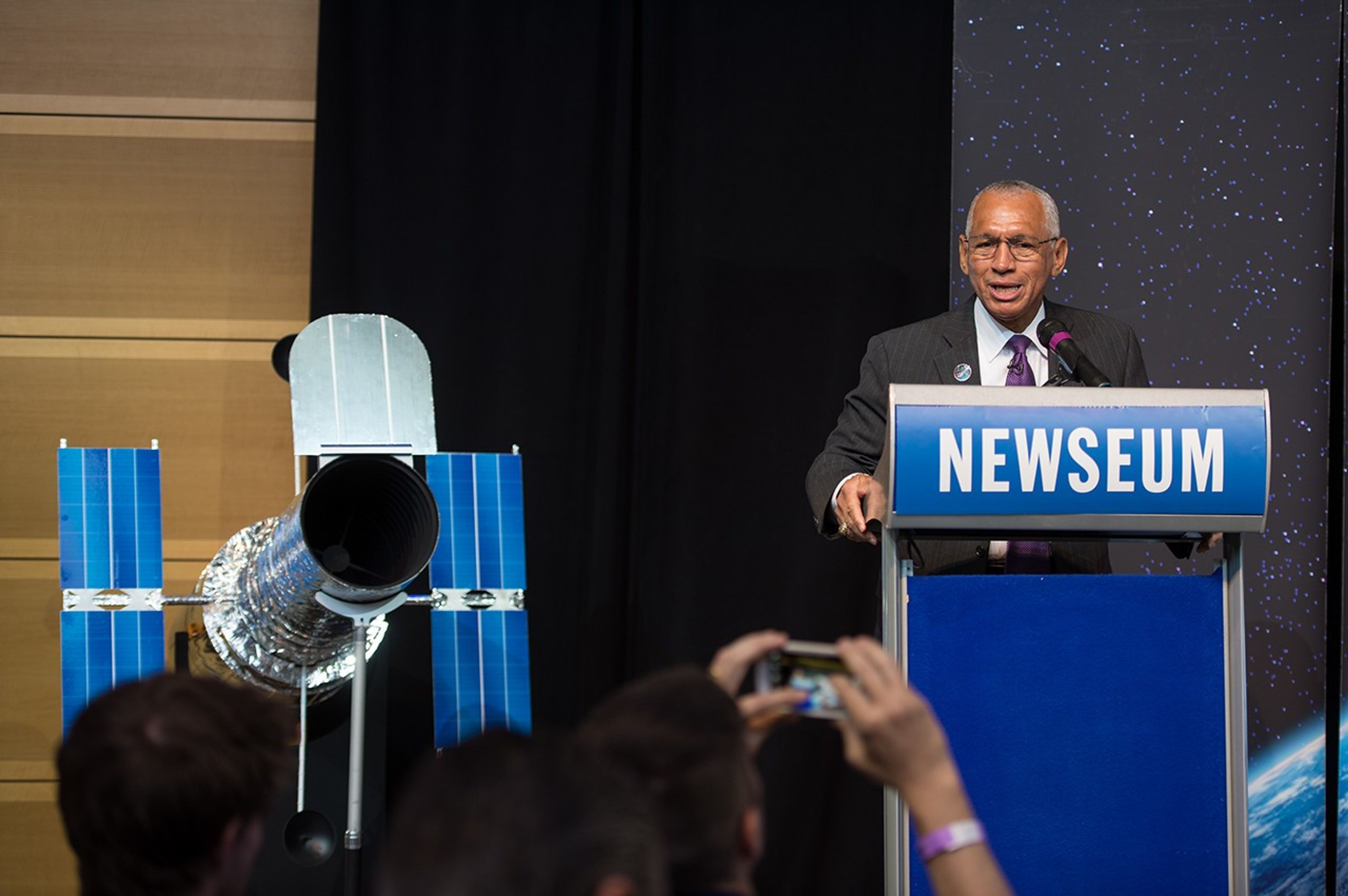 Charlie Bolden stands at the "Newseum" podium on the right side of the picture, addressing the audience; on the left is the Hubble scale model. In the foreground, one of the audience members takes a picture on their phone.