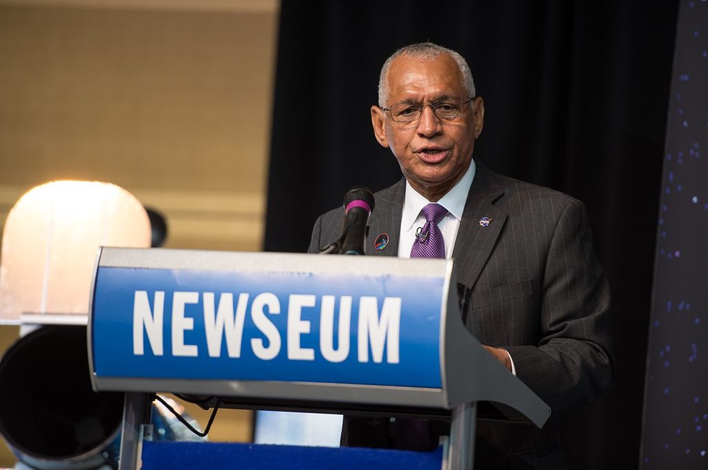 Charlie Bolden stands at the "Newseum" podium, addressing the audience. A scale model of Hubble can be seen in the background on the left.