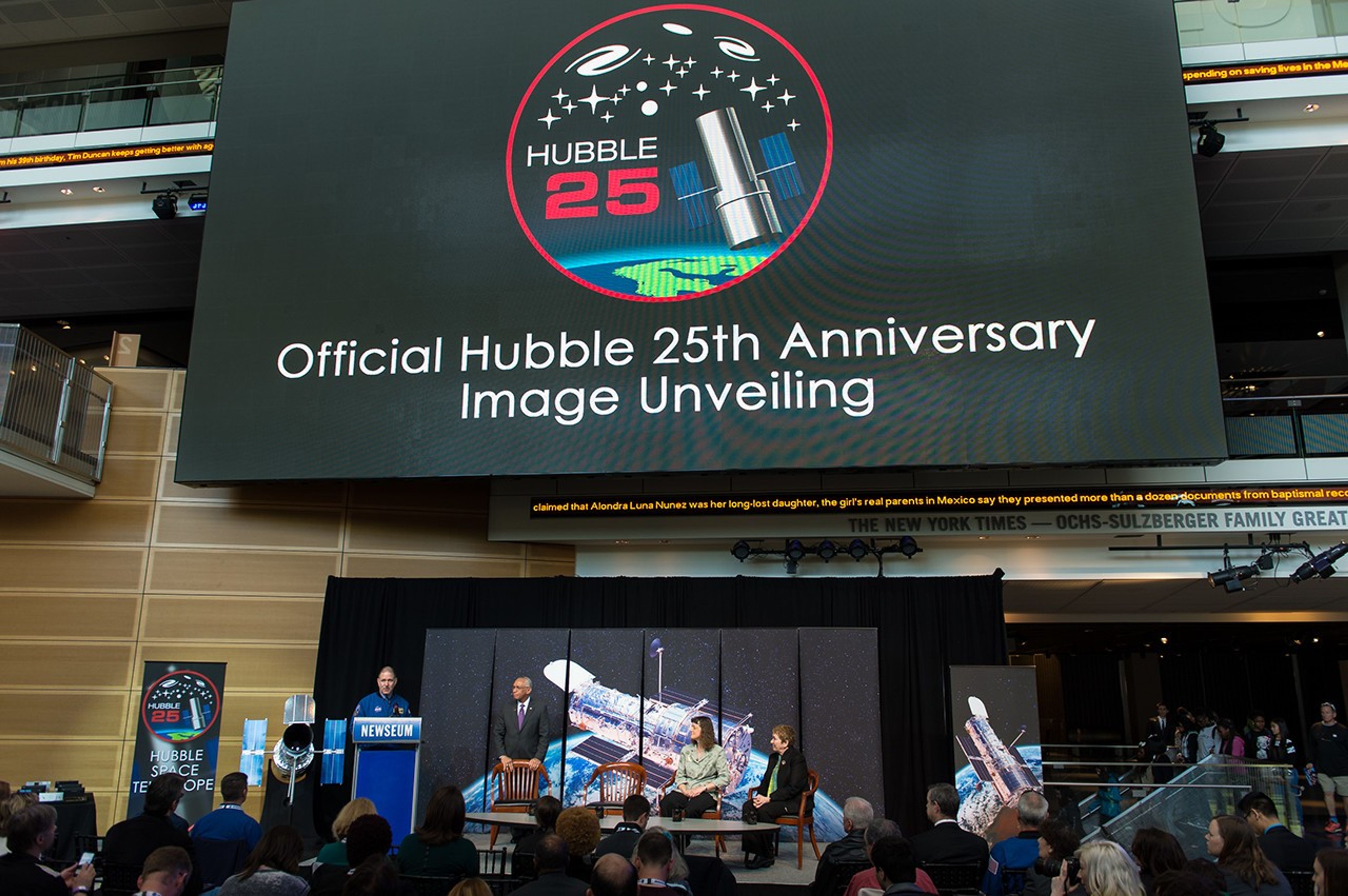 The top half of the image is taken up by a giant screen hanging from the museum's ceiling, projecting a black background with the words "Official Hubble 25th Anniversary Image Unveiling" and the Hubble 25th anniversary logo. In the lower half of the image, Grunsfeld stands at the podium on stage, while Bolden stands by his chair and Drs. Wiseman and Flanagan sit in theirs. Behind them is an image of Hubble over the Earth, left of the stage is the model of Hubble and 25th anniversary banner, and to the right, another banner of Hubble. A large audience is seen watching the presentation.