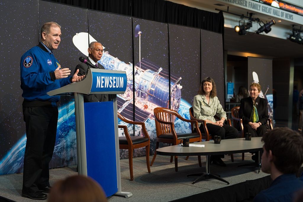Grunsfeld (left) stands at the podium onstage, addressing the audience, while Bolden (center left) stands next to him behind his chair. Dr.s Wiseman (center right) and Flanagan (right) sit listening from their chairs. An image of Hubble over the Earth is behind them.