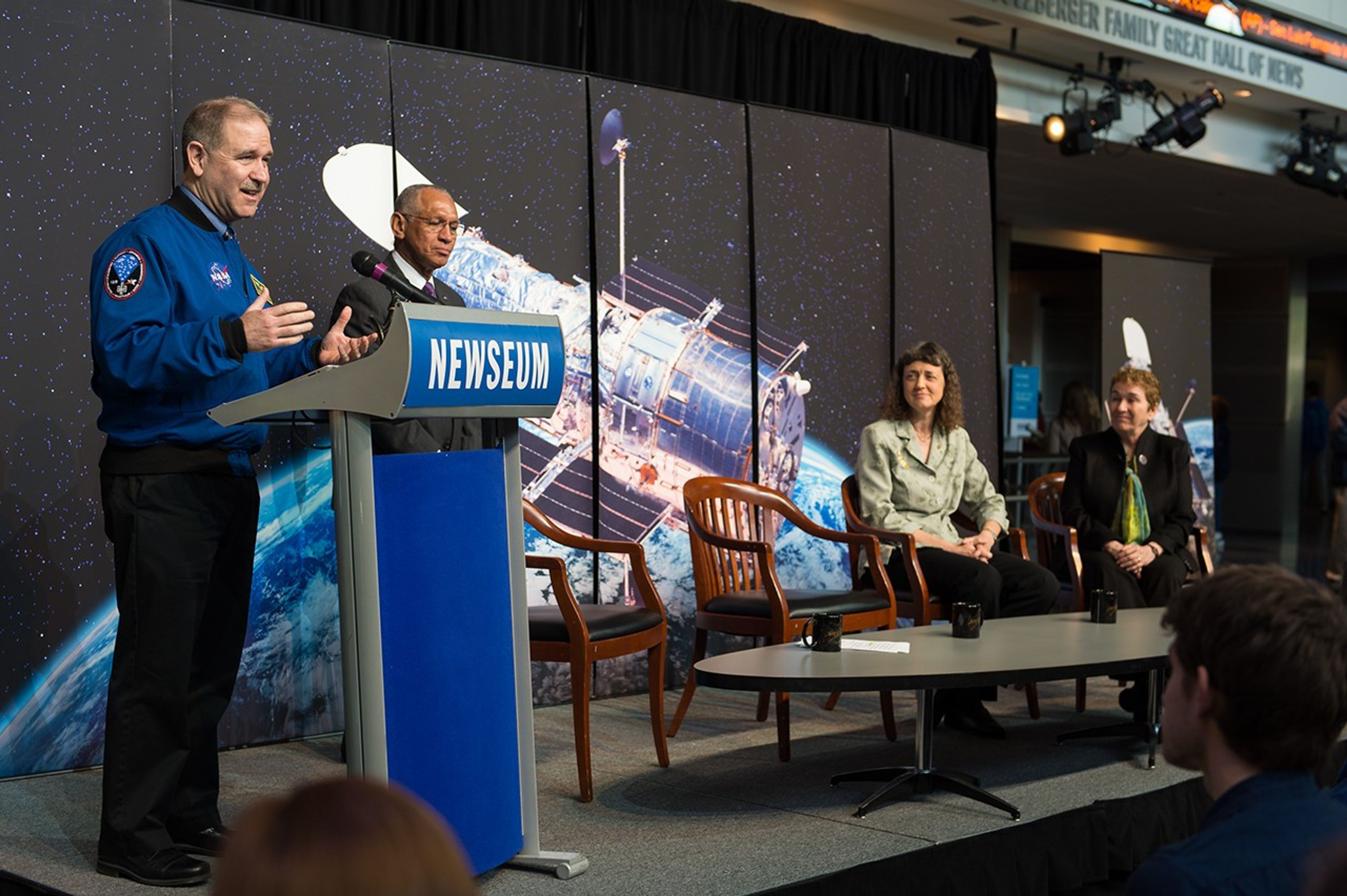 Grunsfeld (left) stands at the podium onstage, addressing the audience, while Bolden (center left) stands next to him behind his chair. Dr.s Wiseman (center right) and Flanagan (right) sit listening from their chairs. An image of Hubble over the Earth is behind them.