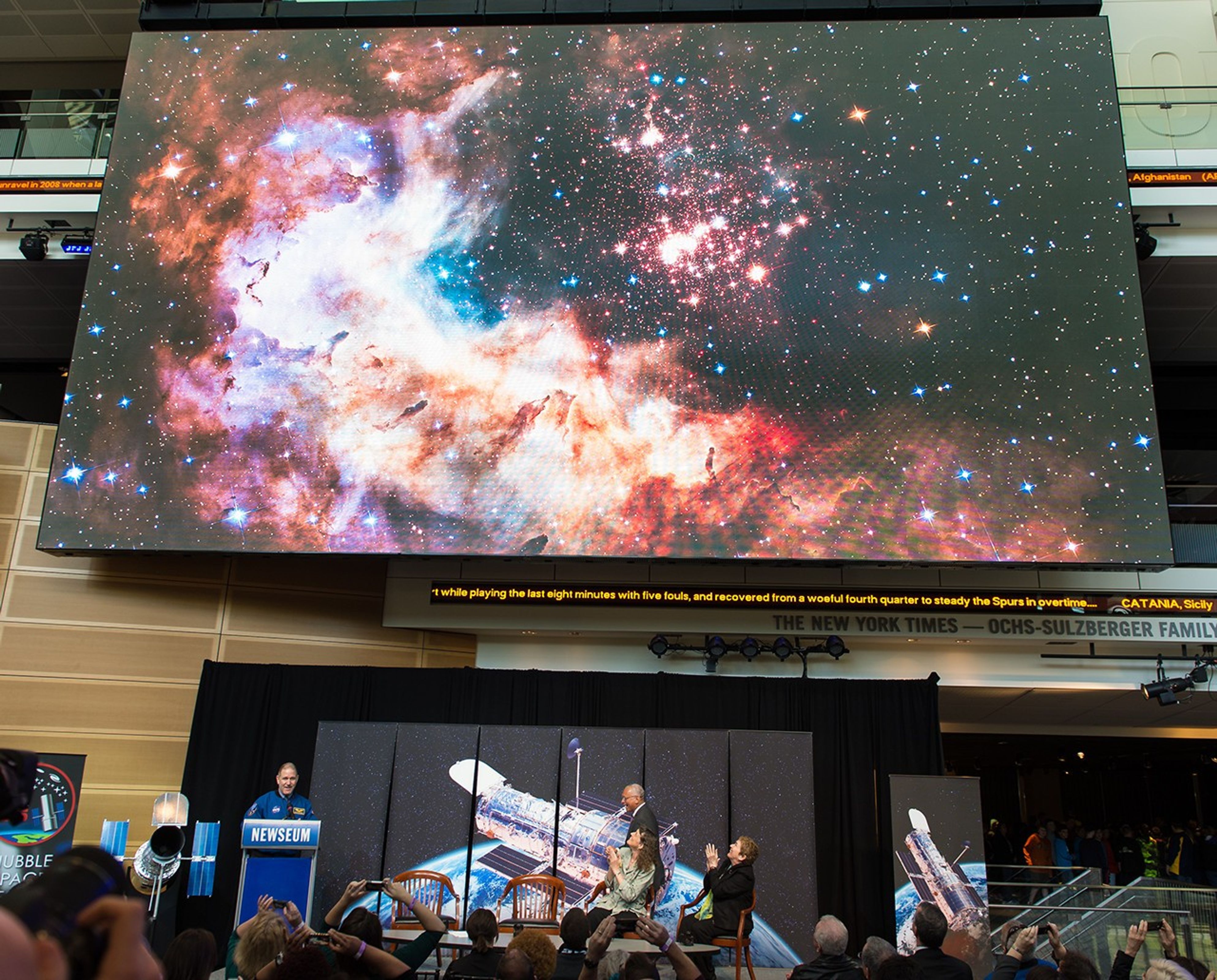 In the top half of the photo, Hubble's image of the star cluster Westerlund 2 is being displayed on the giant half of the screen. In the bottom half, Grunsfeld stands at the podium; Dr.s Flanagan and Wiseman and much of the audience applauds. Several audience members use their phones to take pictures of the screen.