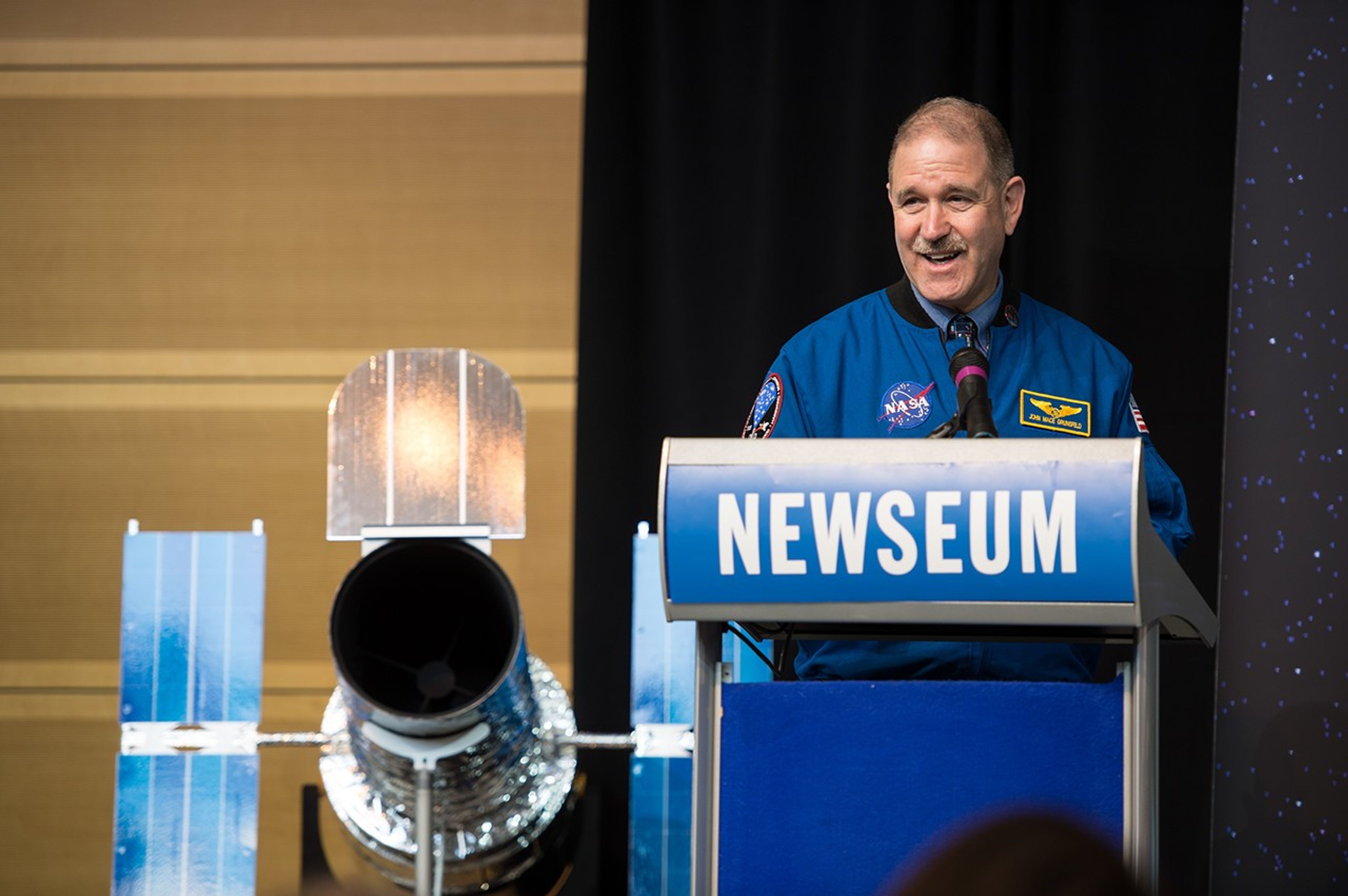 John Grunsfeld stands at the podium, addressing the audience. The scale model of Hubble is visible behind him and to the left.