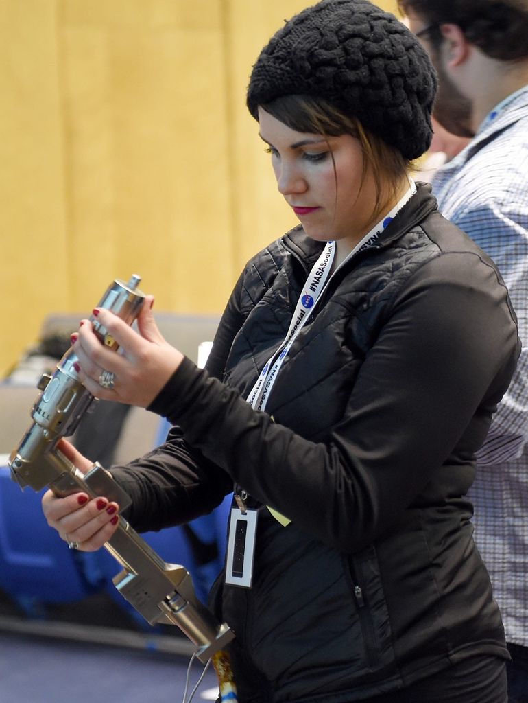 A female visitor holds and inspects a silver L-shaped astronaut tool, the Mini Power Tool.