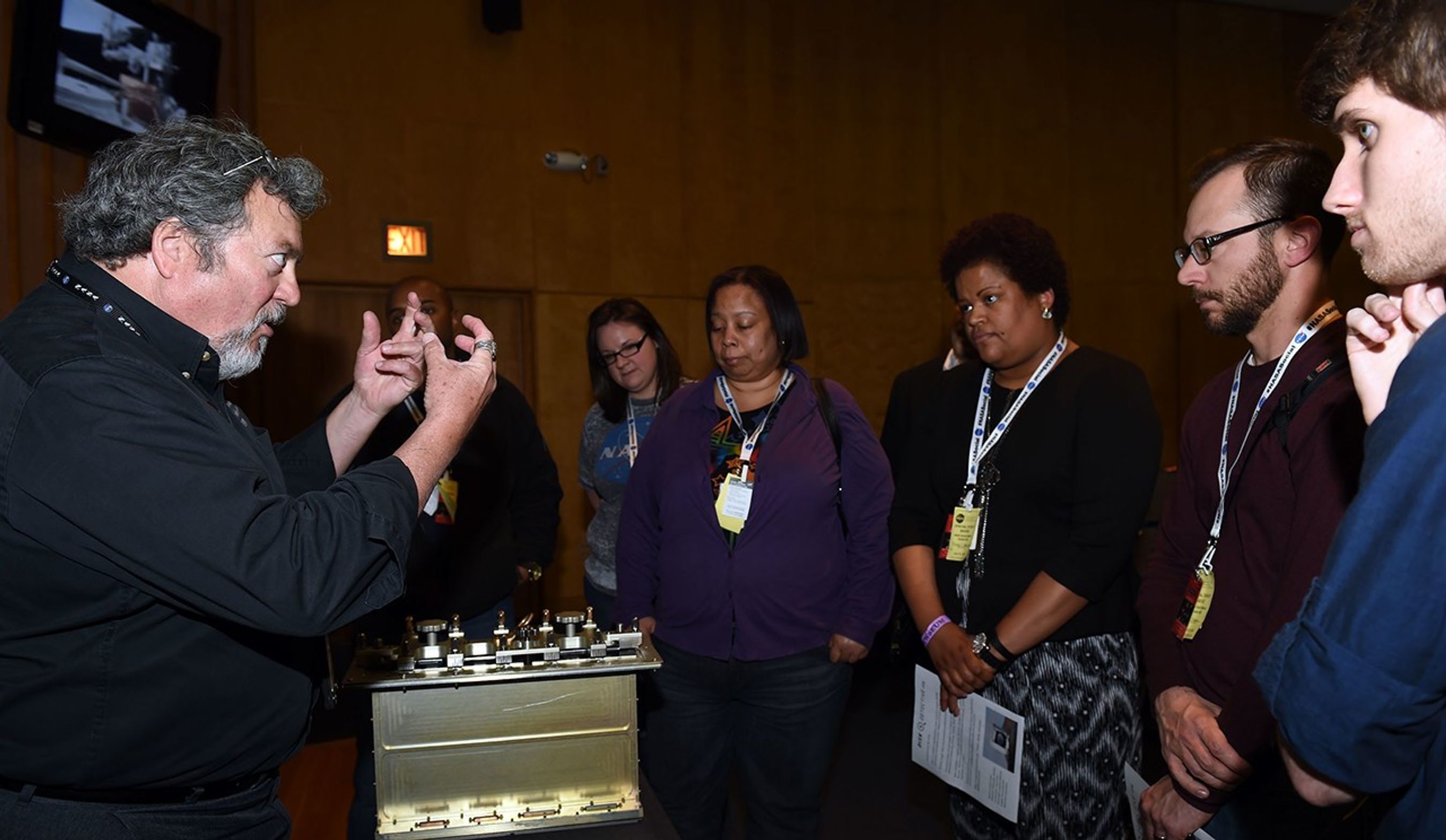 Ed Razac (left) speaks to a group of seven listening visitors (right), with a circuit board module on the table between them.