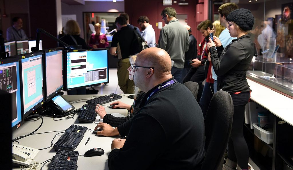 In the foreground, a man and a woman sit side-by-side while working at a series of four computer monitors. Behind them in the background, a large group of visitors listen to a couple more Hubble staff members speaking.