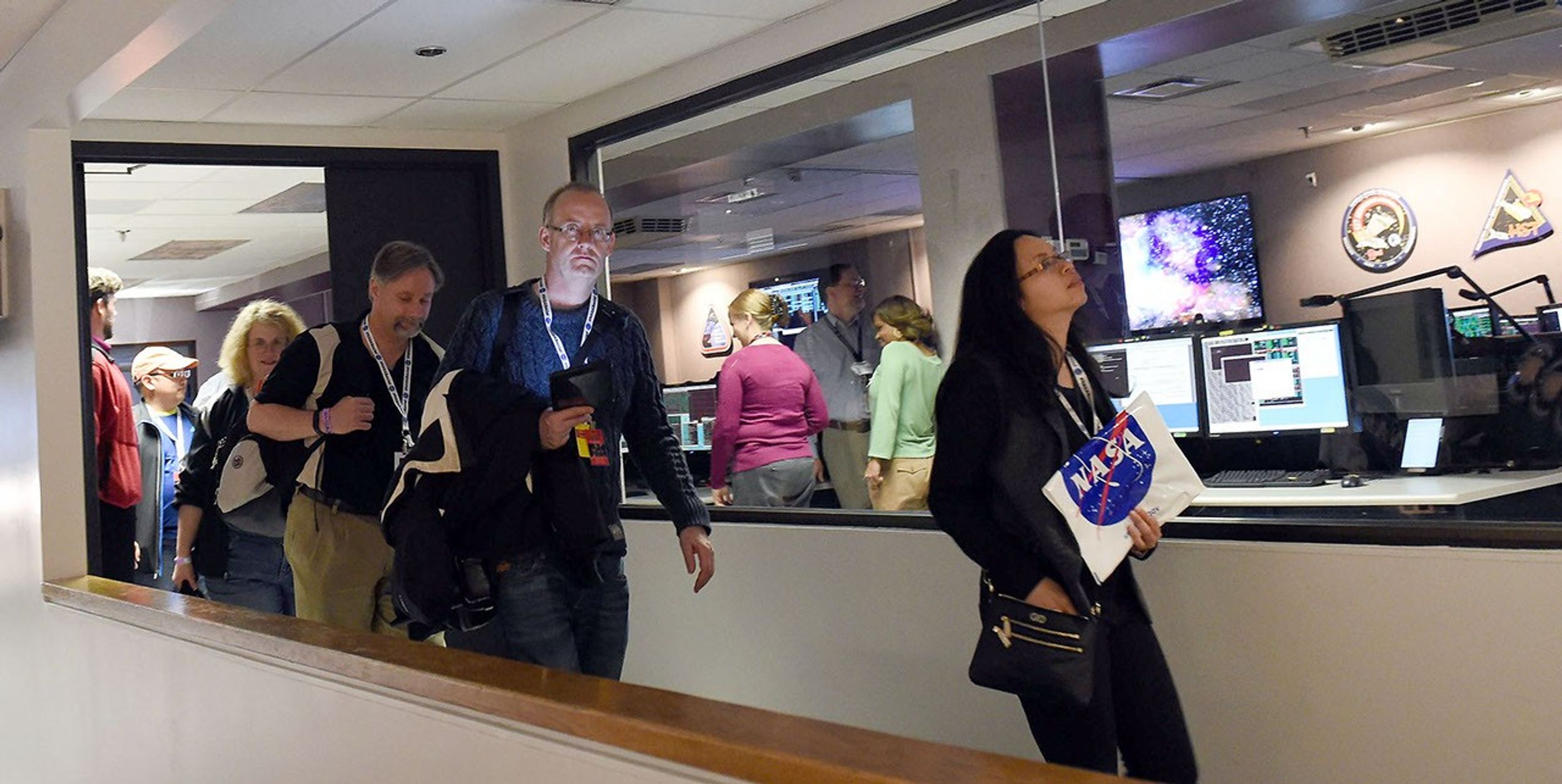 Two women and four men walk to the right of the image out of the Operations Support Room. Three Hubble staff members can be seen in the room in the background through the clear windows.