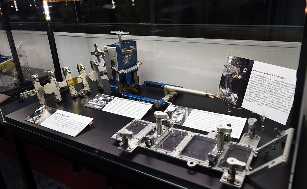 A clear glass case contains three astronaut tools that were used in Hubble servicing missions, with four signs explaining what they are and what they're used for. Another tool from the next case over is visible in the background.