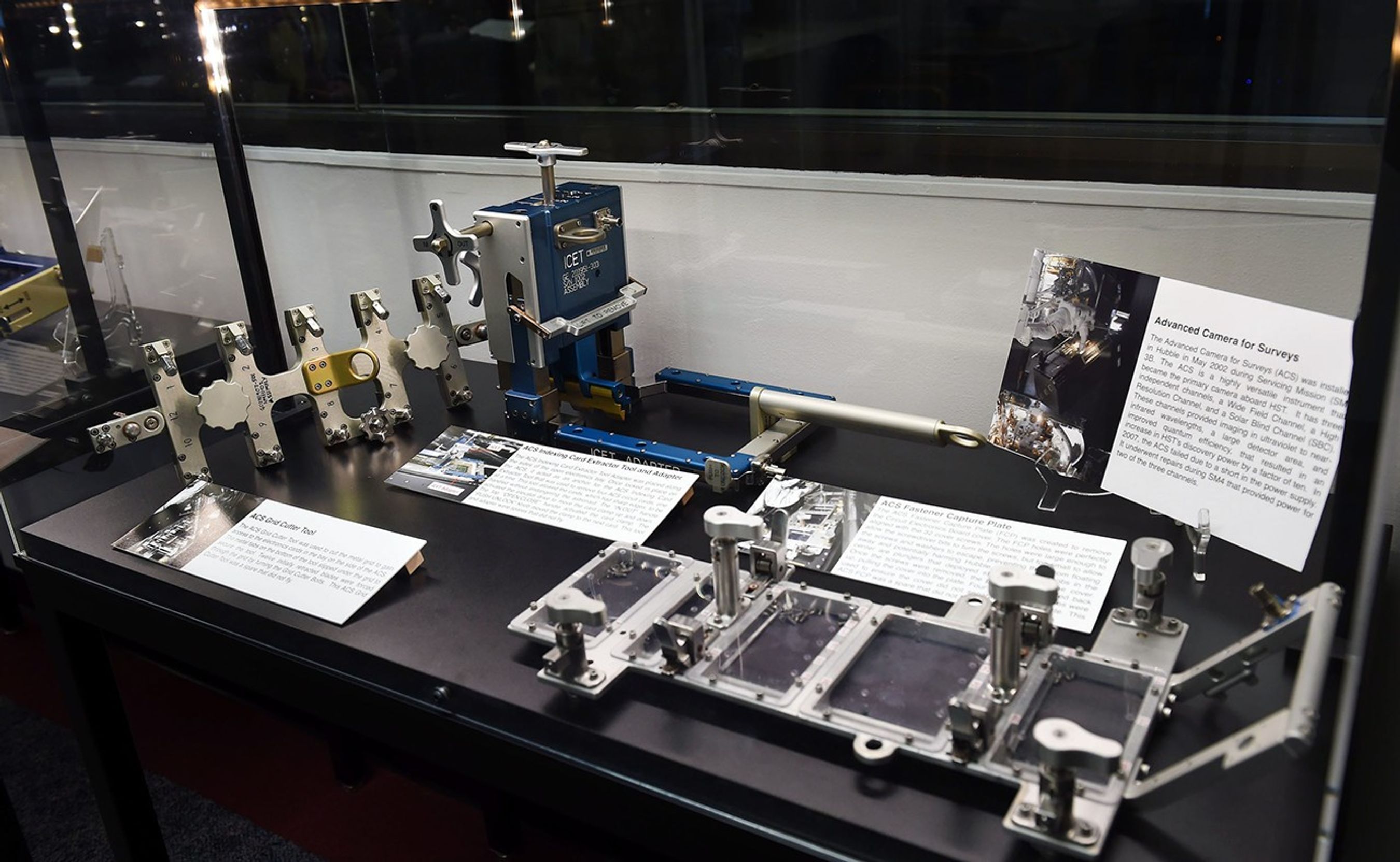 A clear glass case contains three astronaut tools that were used in Hubble servicing missions, with four signs explaining what they are and what they're used for. Another tool from the next case over is visible in the background.