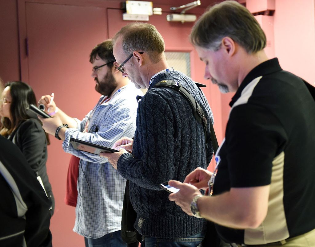 Five visitors face towards the left side of the image as they face an out-of-view speaker and take notes on phones or tablets.