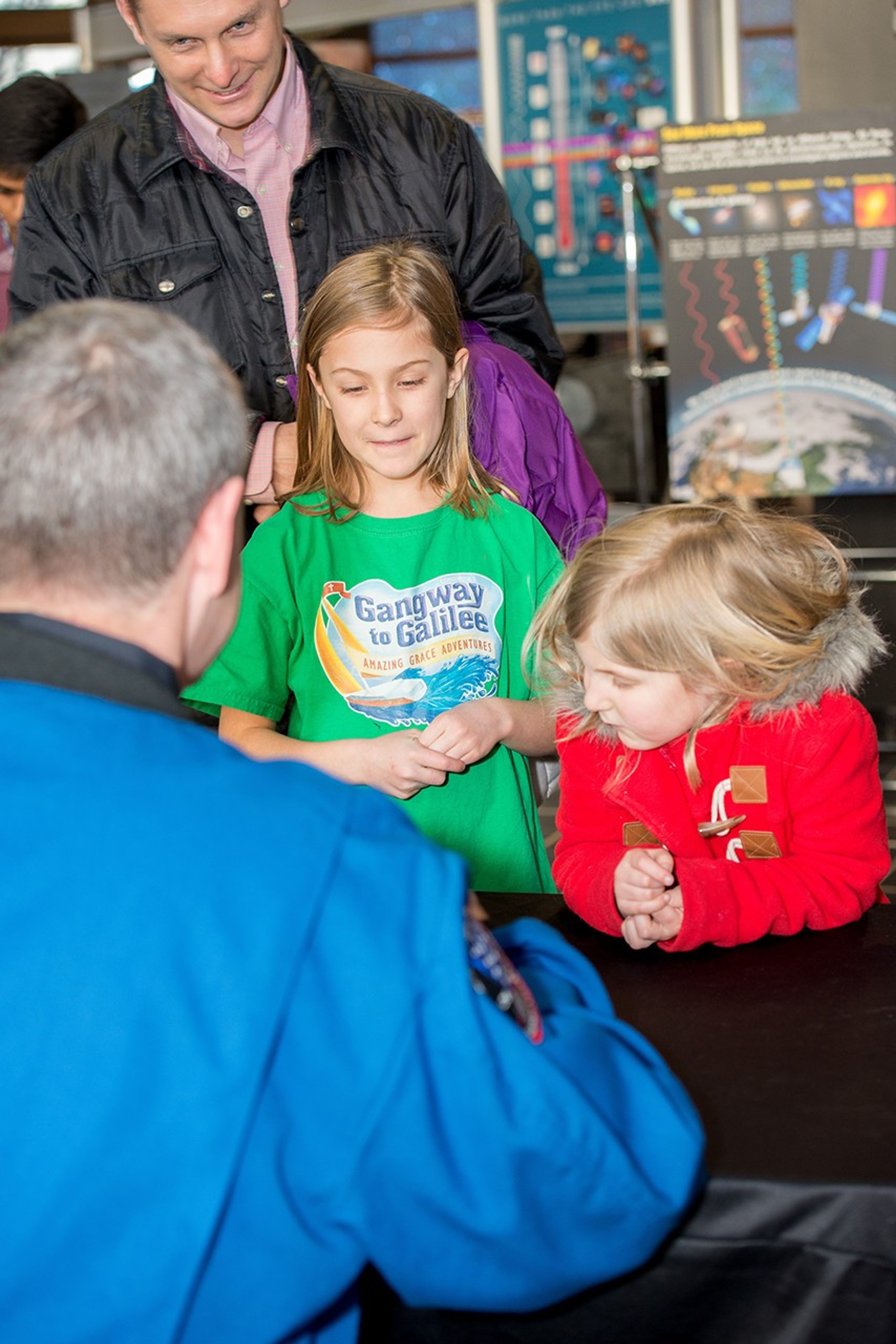 In the foreground, astronaut Scott Altman sits behind a table and faces away from the camera toward two young girls and the male adult with them on the other side of the table.